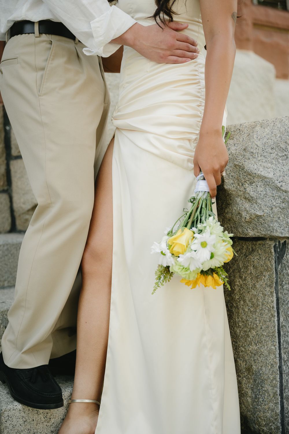 Bride in ivory satin gown holding yellow and white bouquet stands with groom in beige trousers on stone steps.