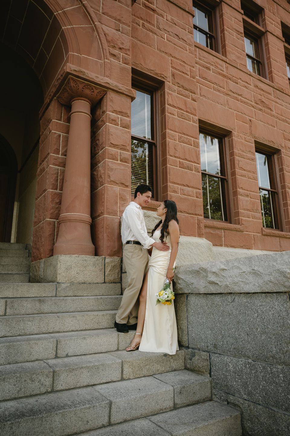 Bride in white gown and groom share a romantic moment on stone steps of a historic sandstone building.