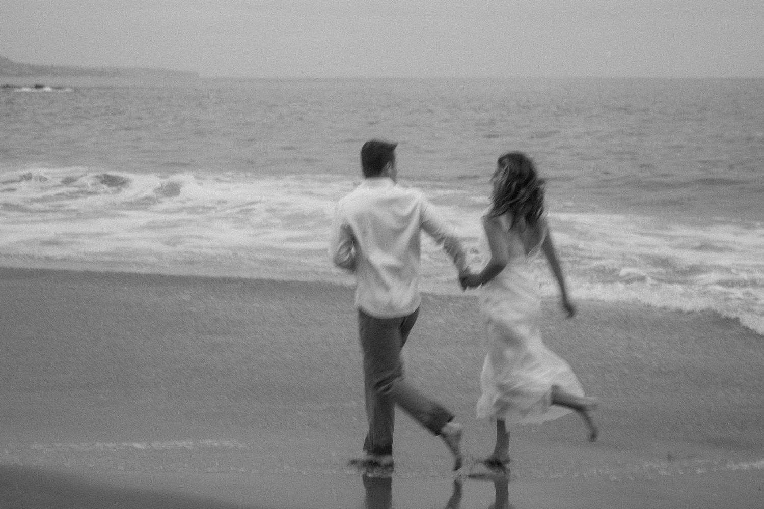 Black and white photo of a couple holding hands and running playfully along a sandy beach with ocean waves.