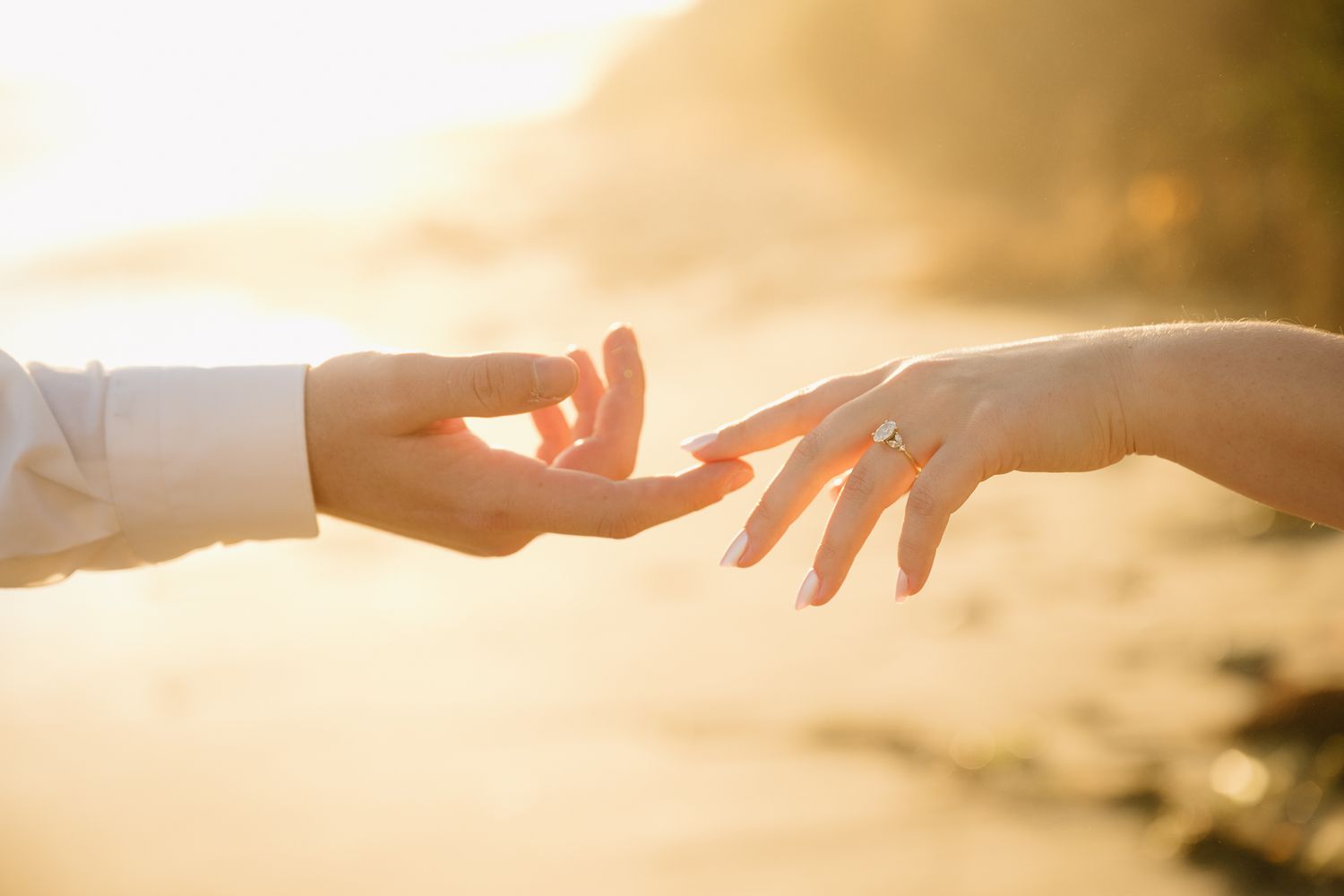 Two hands reaching toward each other at golden sunset beach, woman wearing engagement ring, romantic couple moment.