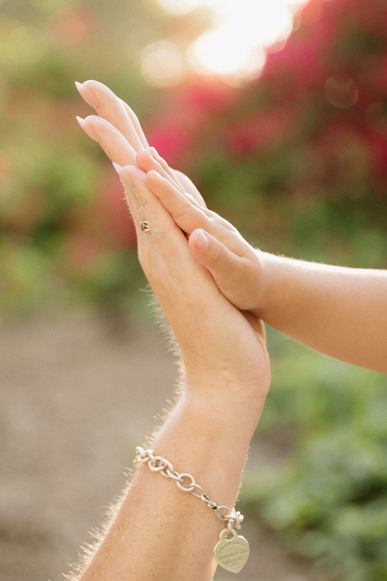 Two hands pressed together palm-to-palm outdoors, one wearing a silver charm bracelet, with a soft bokeh garden background.