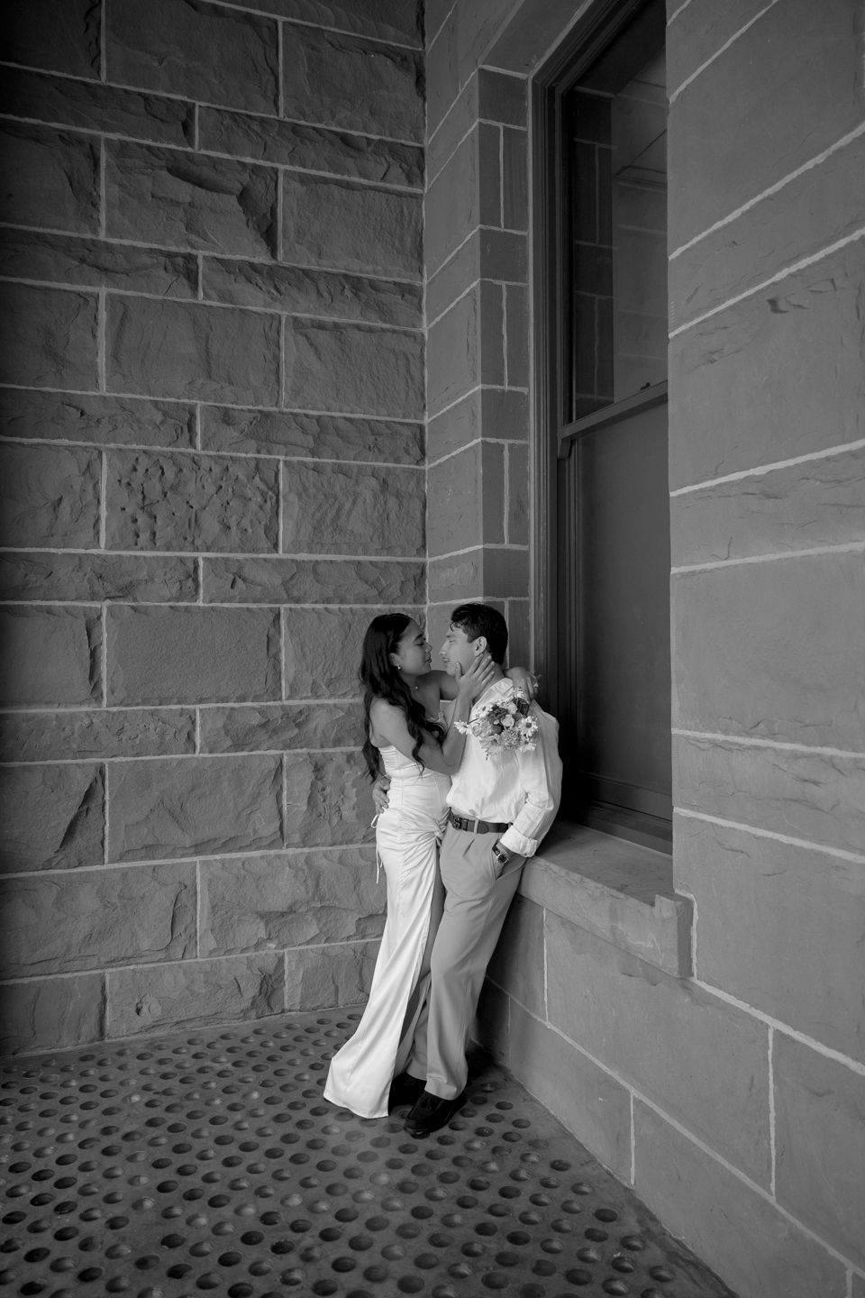Black and white photo of couple sharing romantic moment by stone building window.