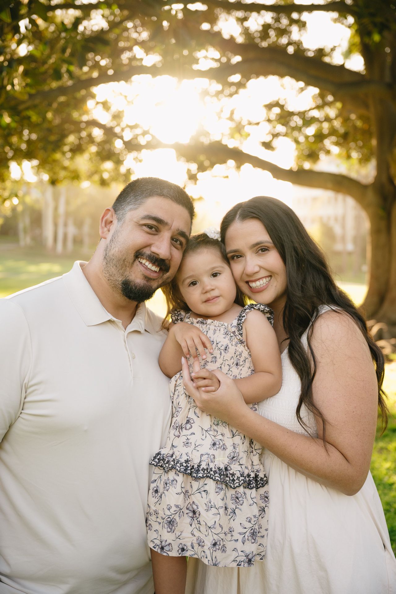 A smiling family of three poses outdoors under a sunlit tree, with parents holding their young daughter in a floral dress.