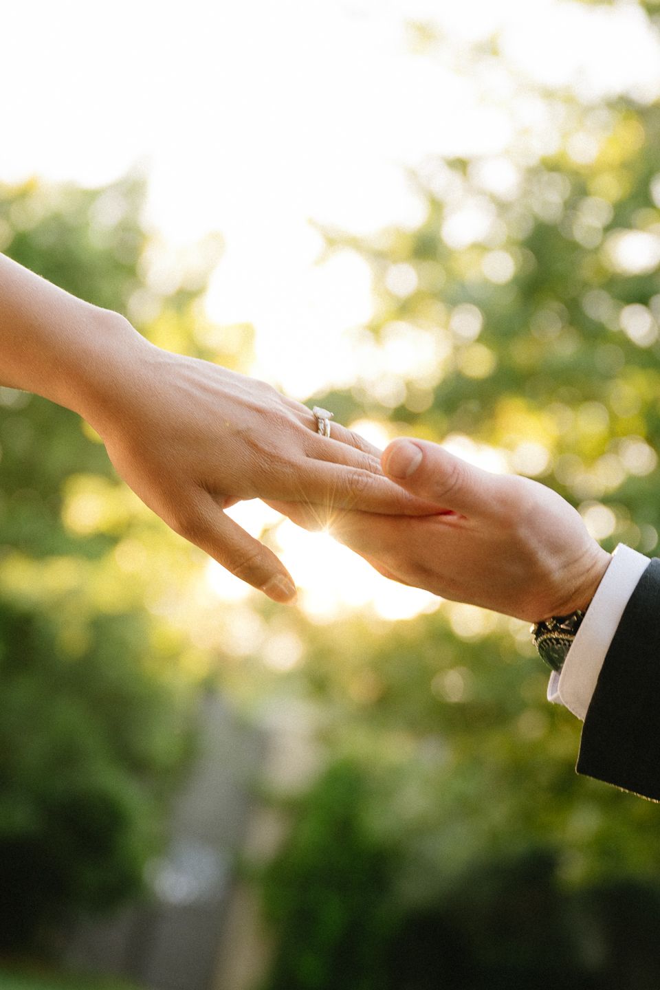 Bride and groom holding hands outdoors, engagement ring visible, sunlit green trees in background.