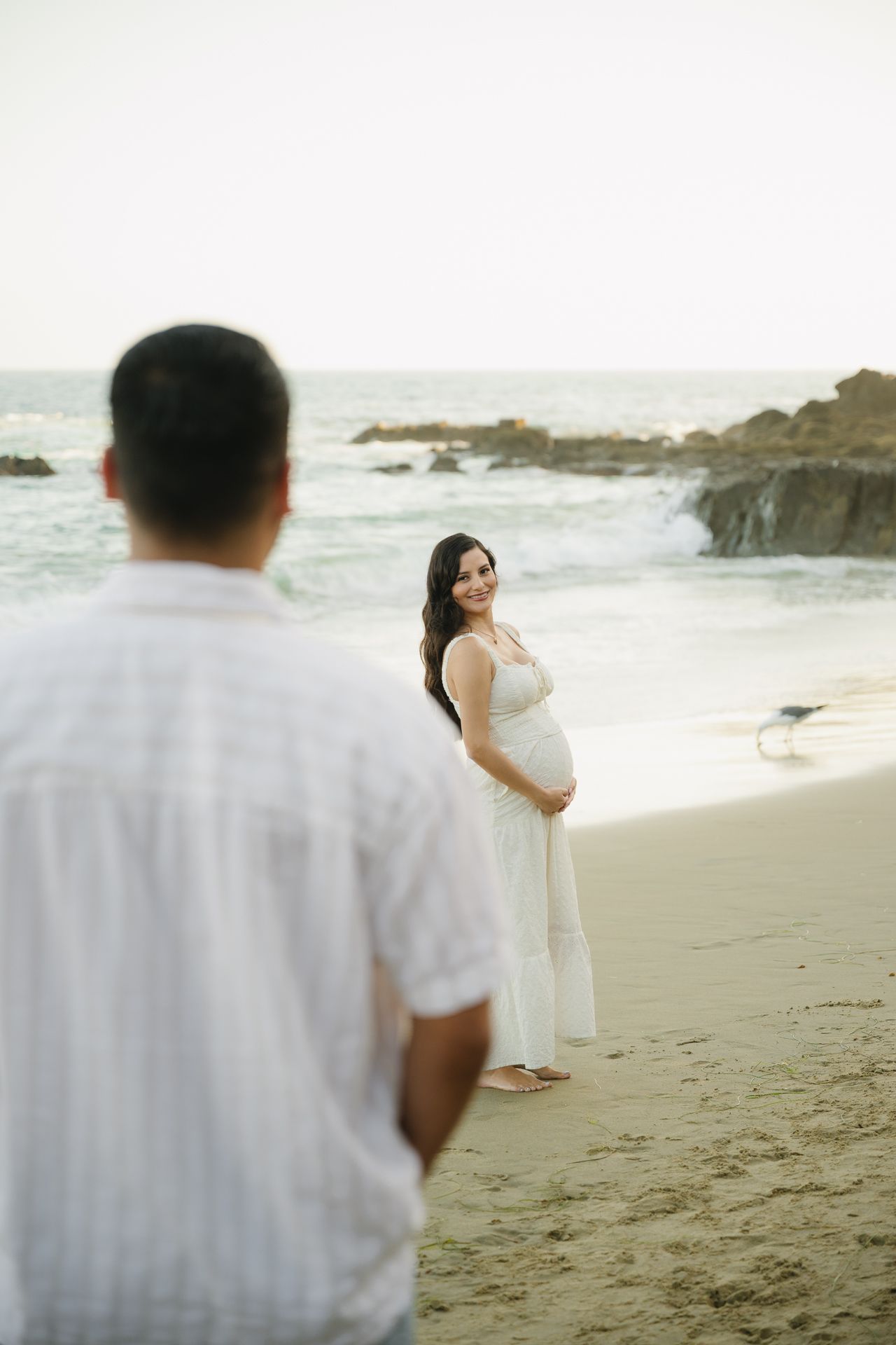 Pregnant woman in white dress smiling on beach while man in white shirt stands in foreground.