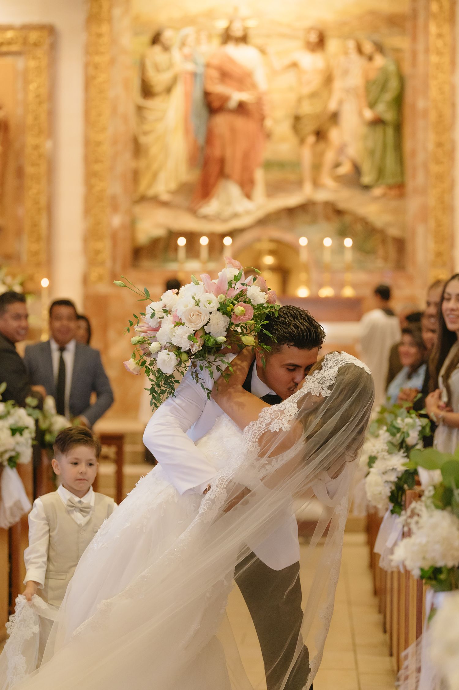 Bride and groom share first kiss at ornate church altar, bride holding lush white and pink bouquet.
