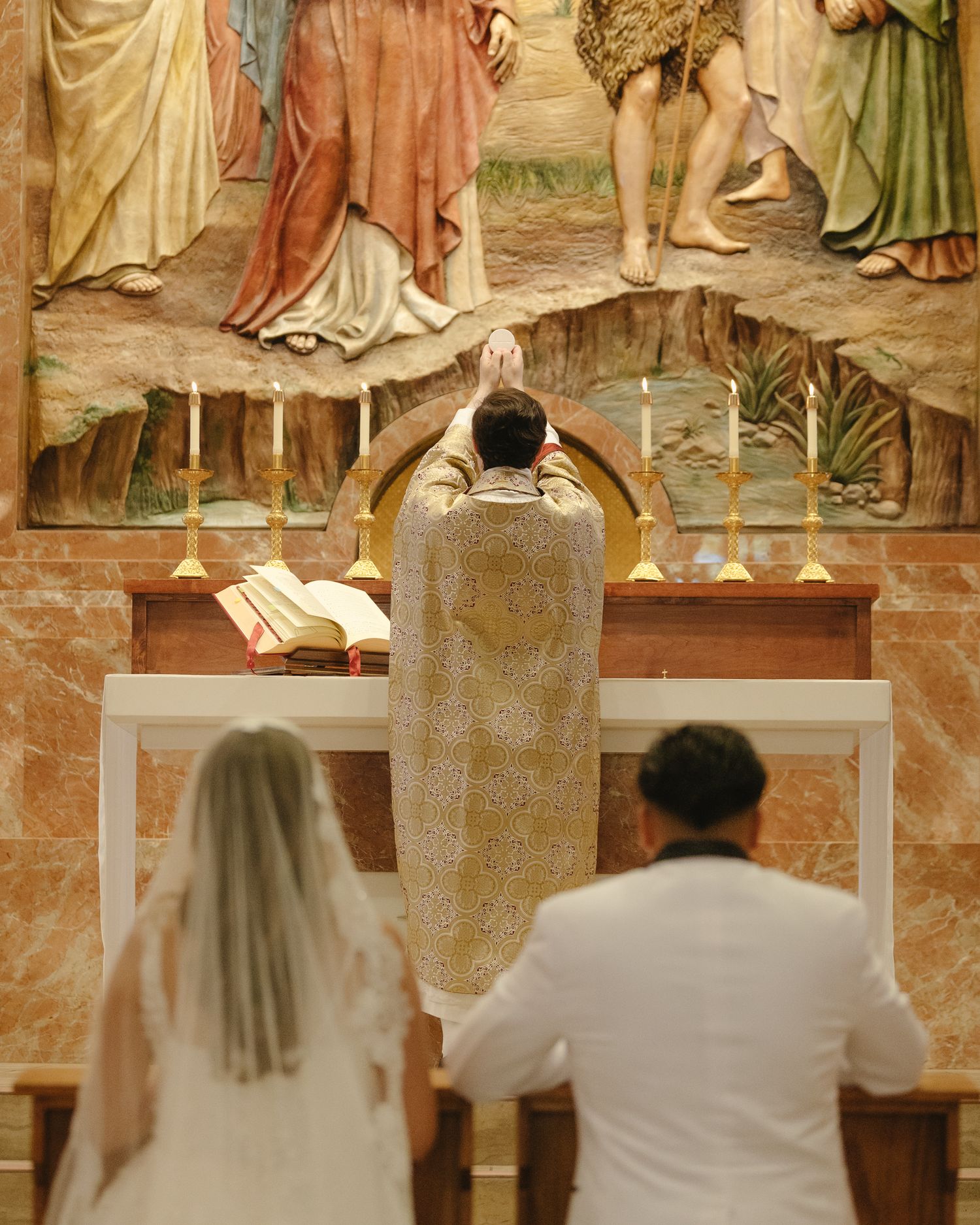 Bride and groom kneel before priest at ornate church altar during Catholic wedding ceremony with religious mural backdrop.