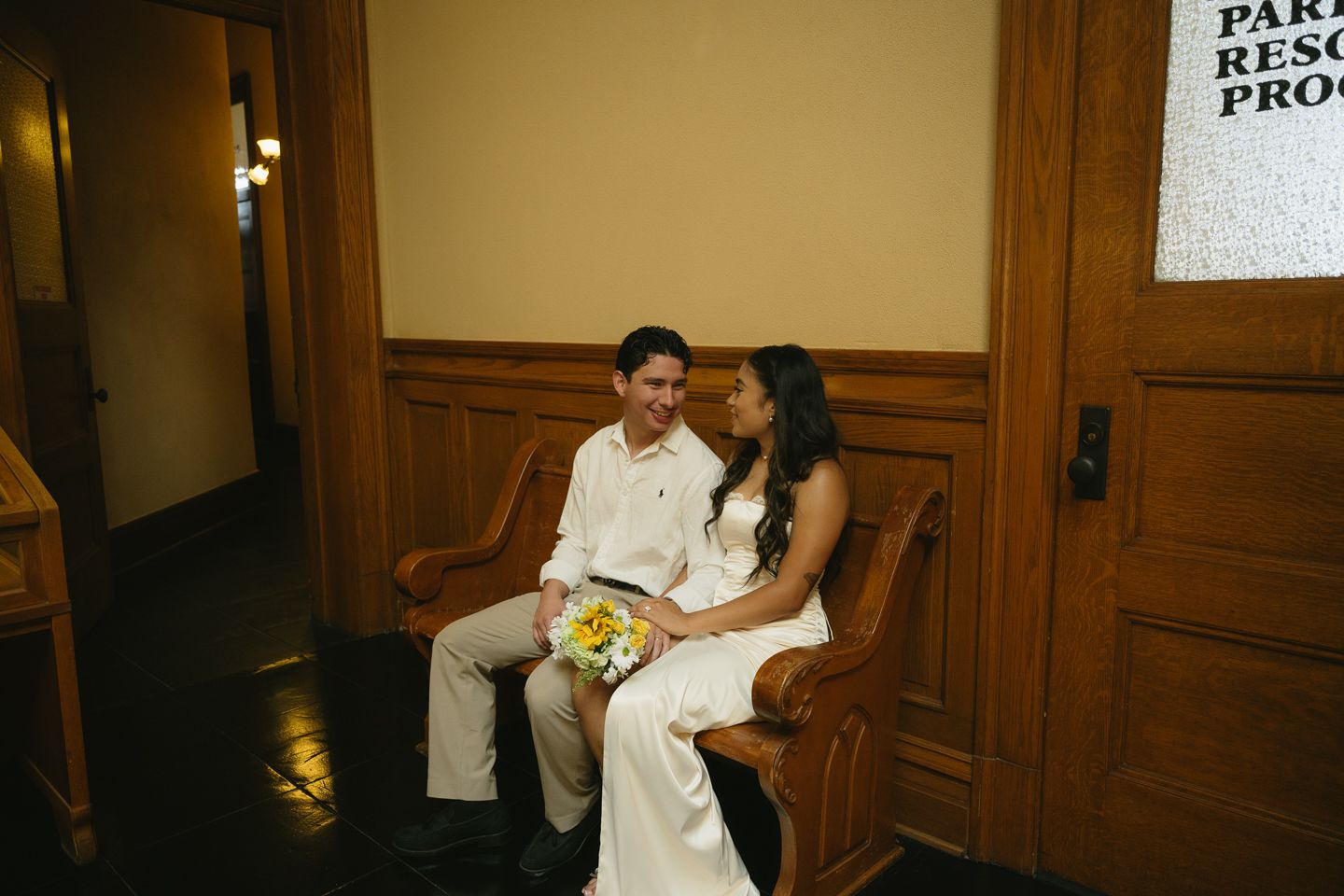 A bride and groom sit together on a wooden bench, smiling, as the bride holds a yellow bouquet in a warmly lit hall.