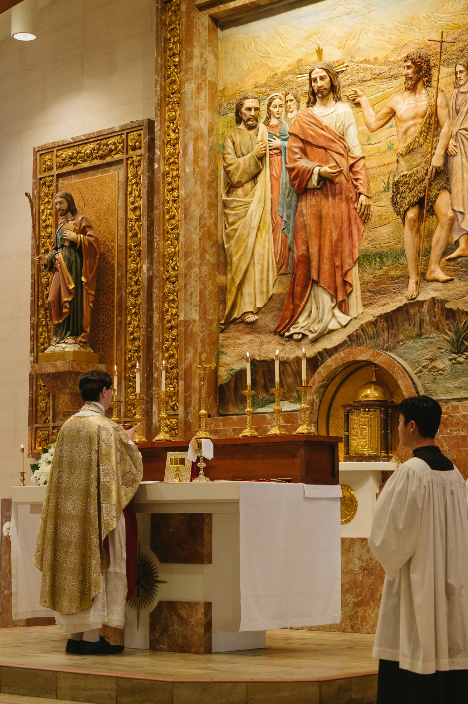 Catholic priest and altar server stand before ornate altar with large religious mural depicting Jesus, Mary, and saints in a church.
