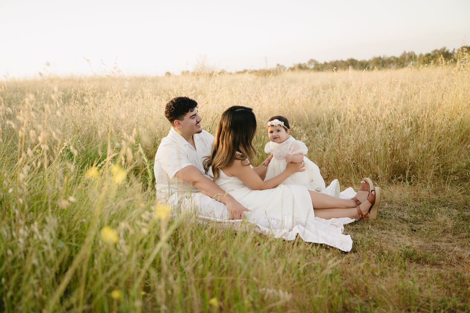 Family sitting in golden field, parents smiling at baby in white dress during sunny outdoor photo session.