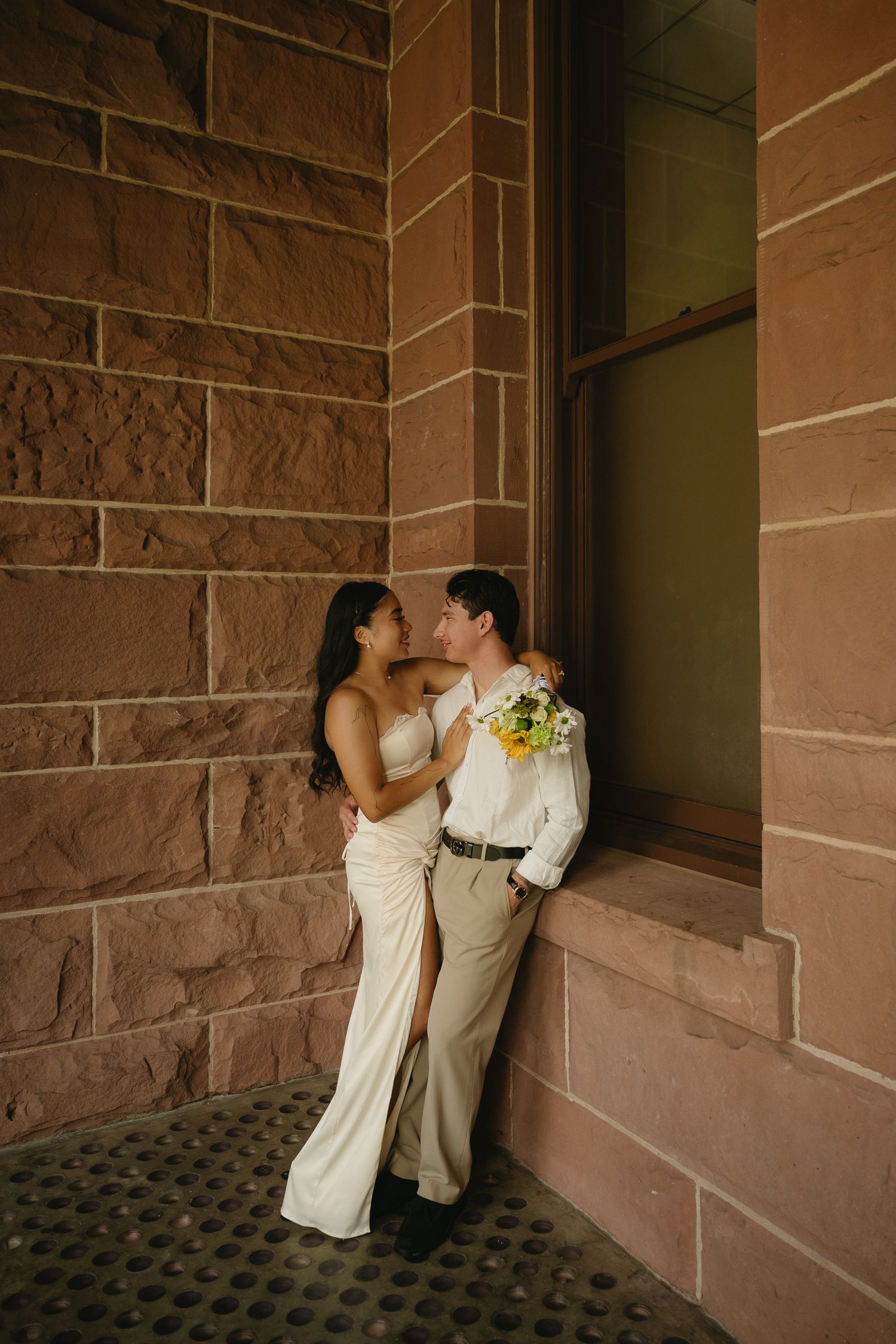 A couple in wedding attire embraces near a sandstone wall, the bride in a white gown holding a yellow bouquet.