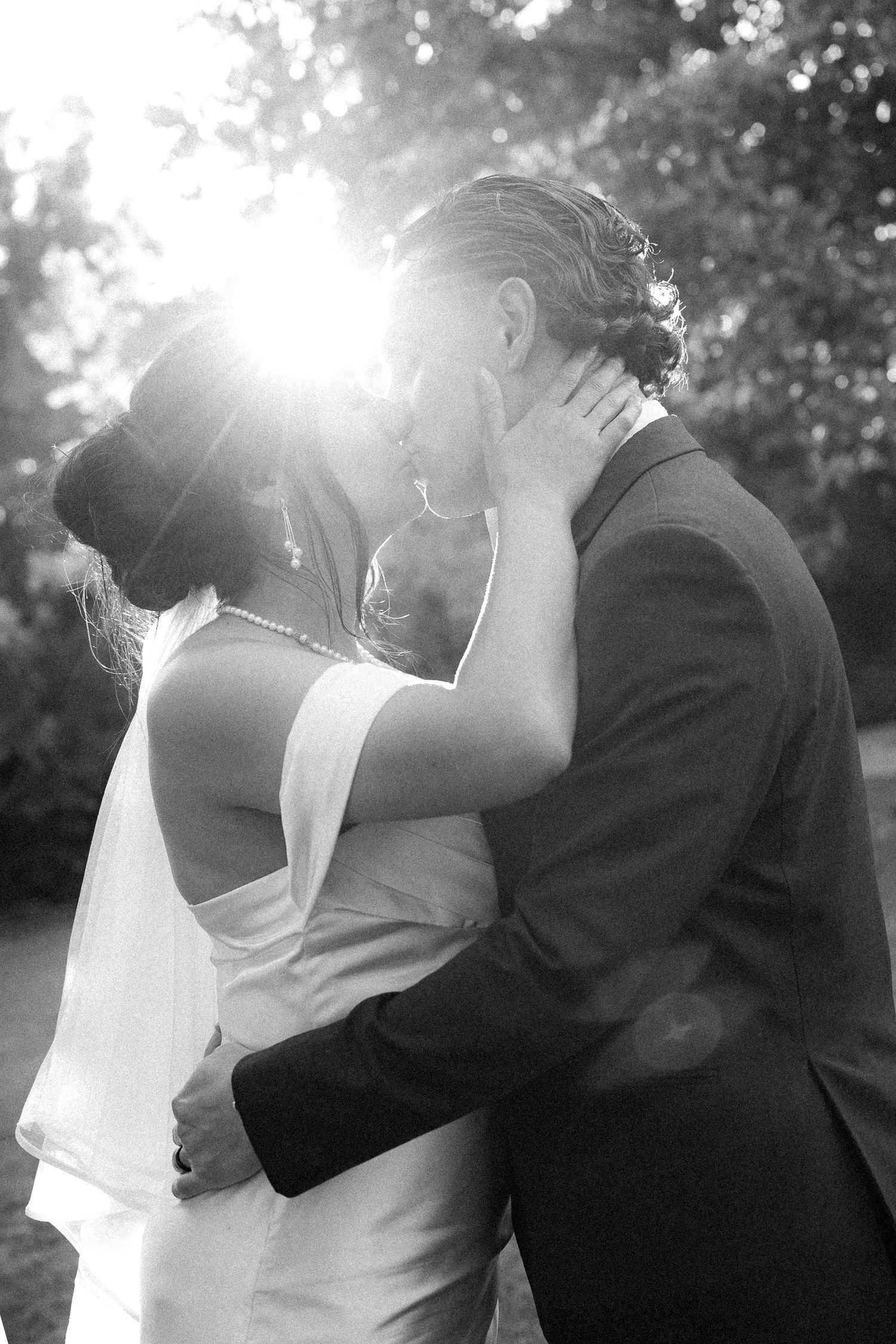 Black and white wedding photo of bride and groom sharing a romantic kiss backlit by sunlight outdoors.