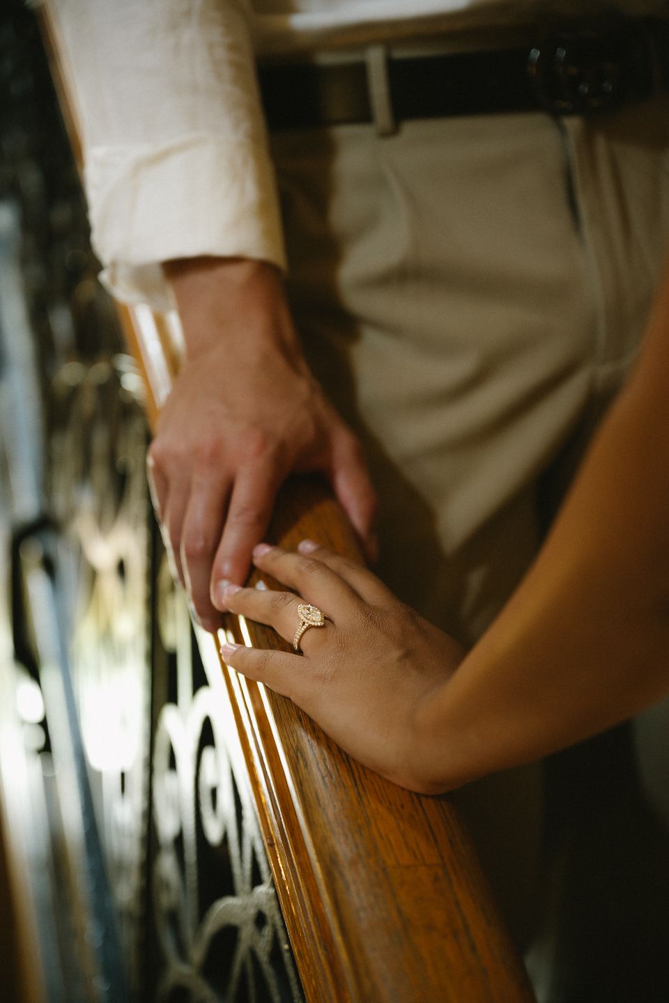 Two hands touching on a wooden railing, one wearing a gold ring, with a man in beige trousers standing nearby.