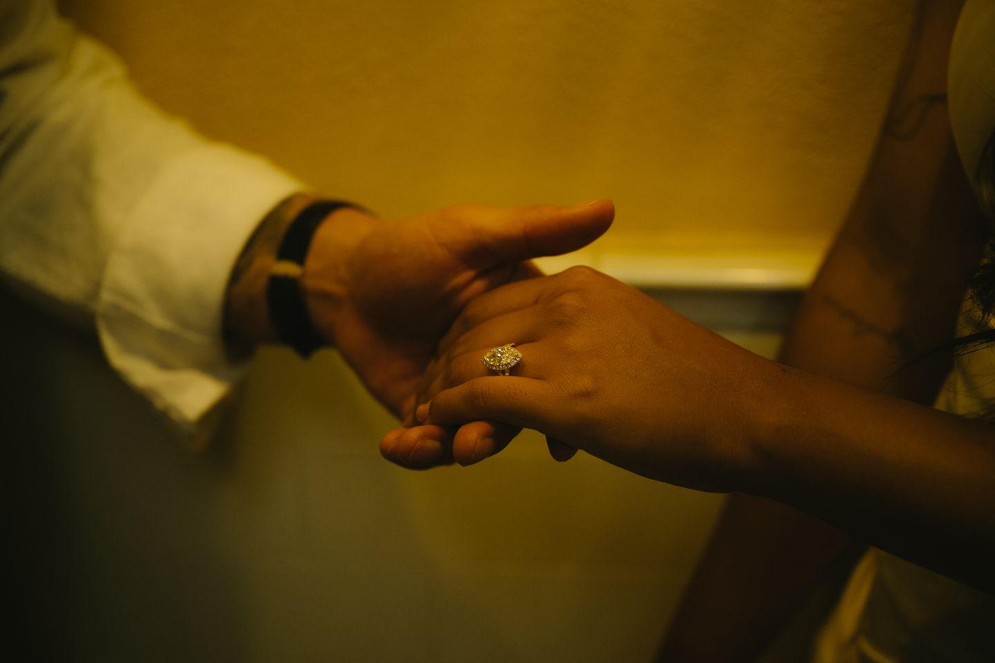 Close-up of two hands gently touching, one wearing a diamond engagement ring, bathed in warm golden light.