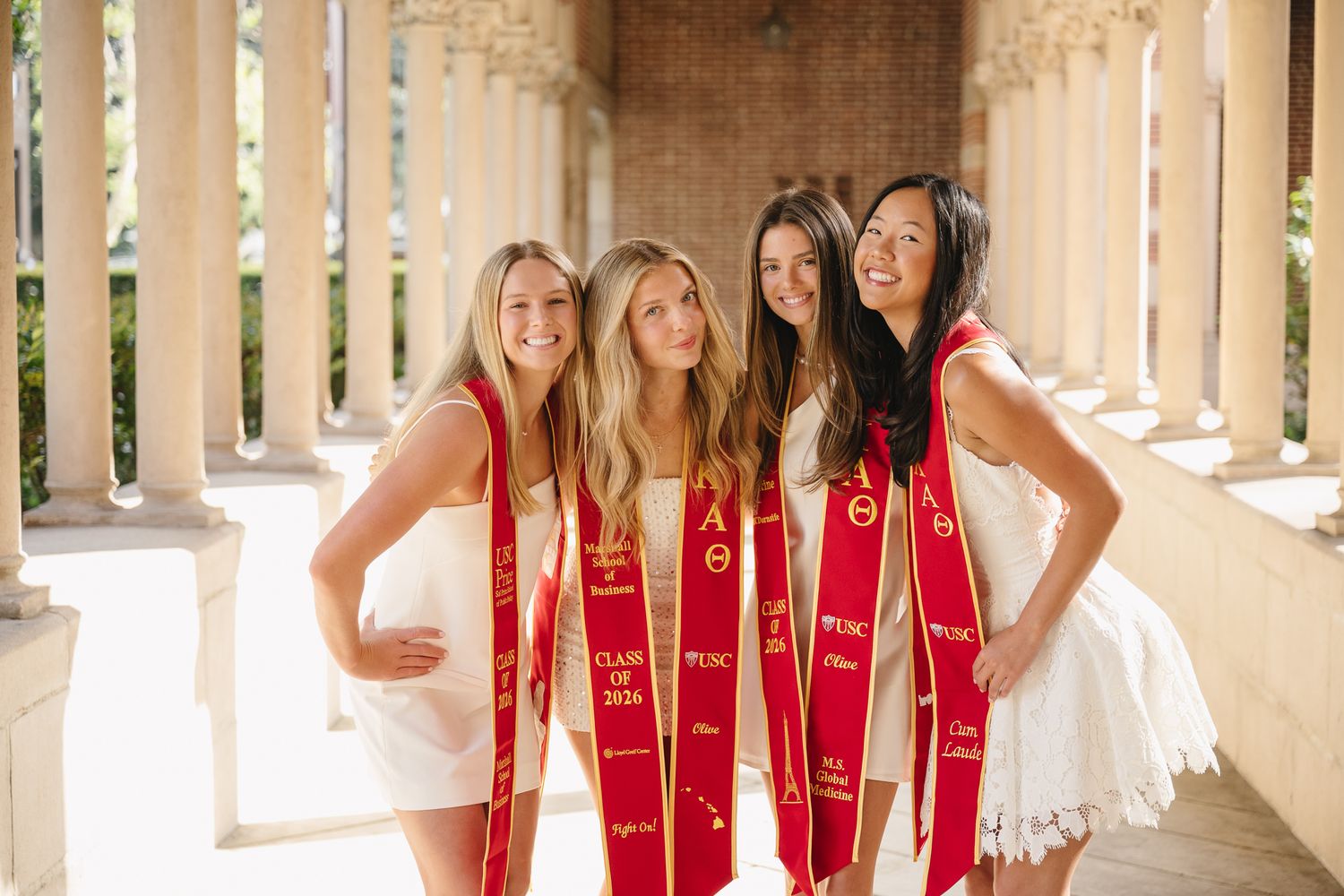Four women in white dresses wearing red graduation sashes pose together in a sunlit colonnade.