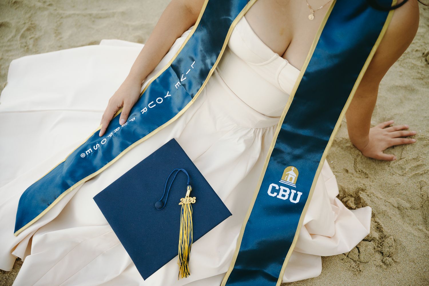 CBU graduation cap with gold tassel and blue sash on white dress at beach ceremony.