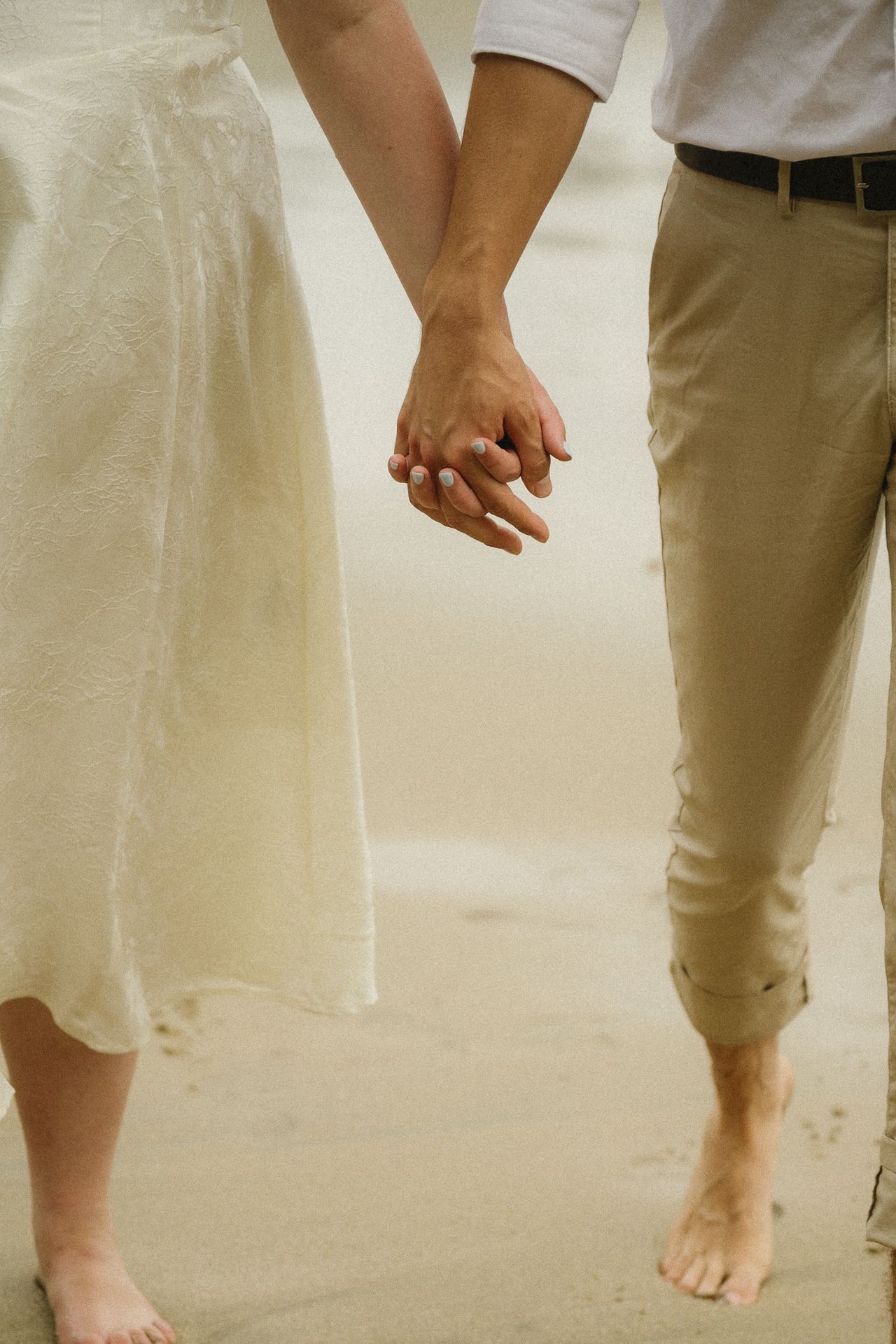 Couple holding hands on sandy beach, woman in white dress and man in beige pants, barefoot romantic moment.