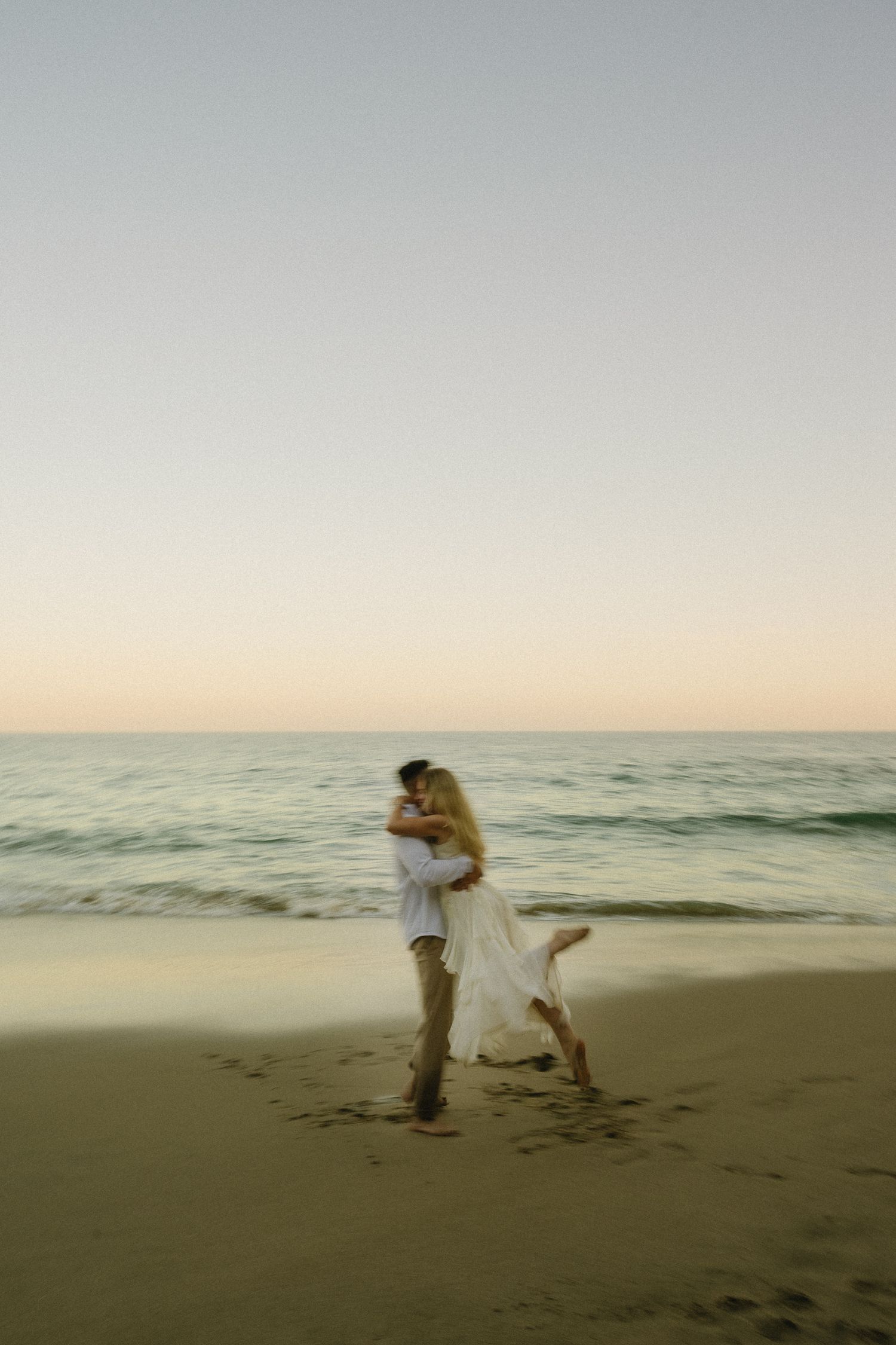 Couple embracing on a Laguna Beach at Crescent Bay at sunset, waves gently rolling in behind them under a pastel sky.