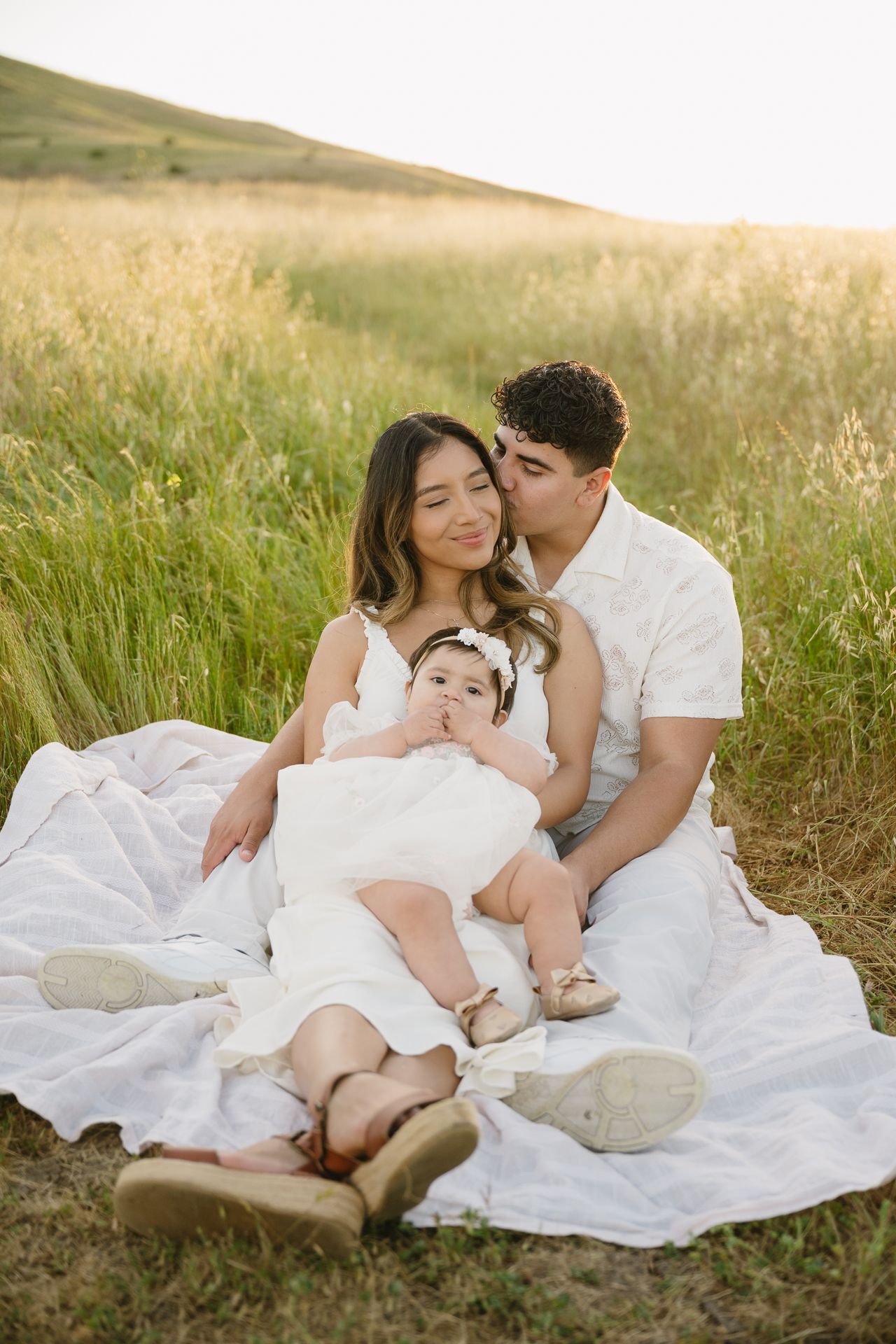 A young family in white outfits sits on a blanket in a sunlit field, parents lovingly holding their baby girl.