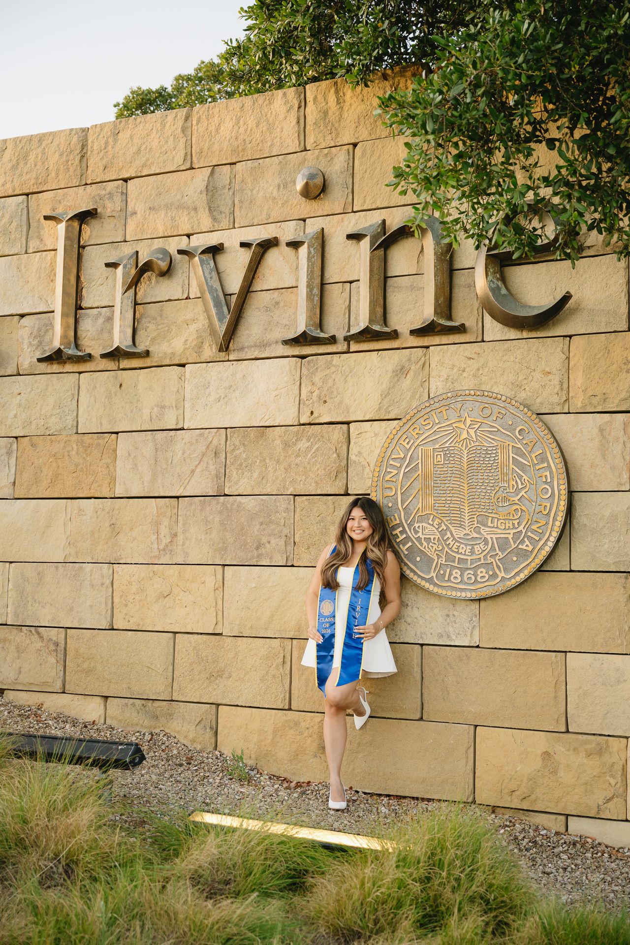 Graduate poses in front of UC Irvine stone wall sign wearing white dress and blue graduation stole.