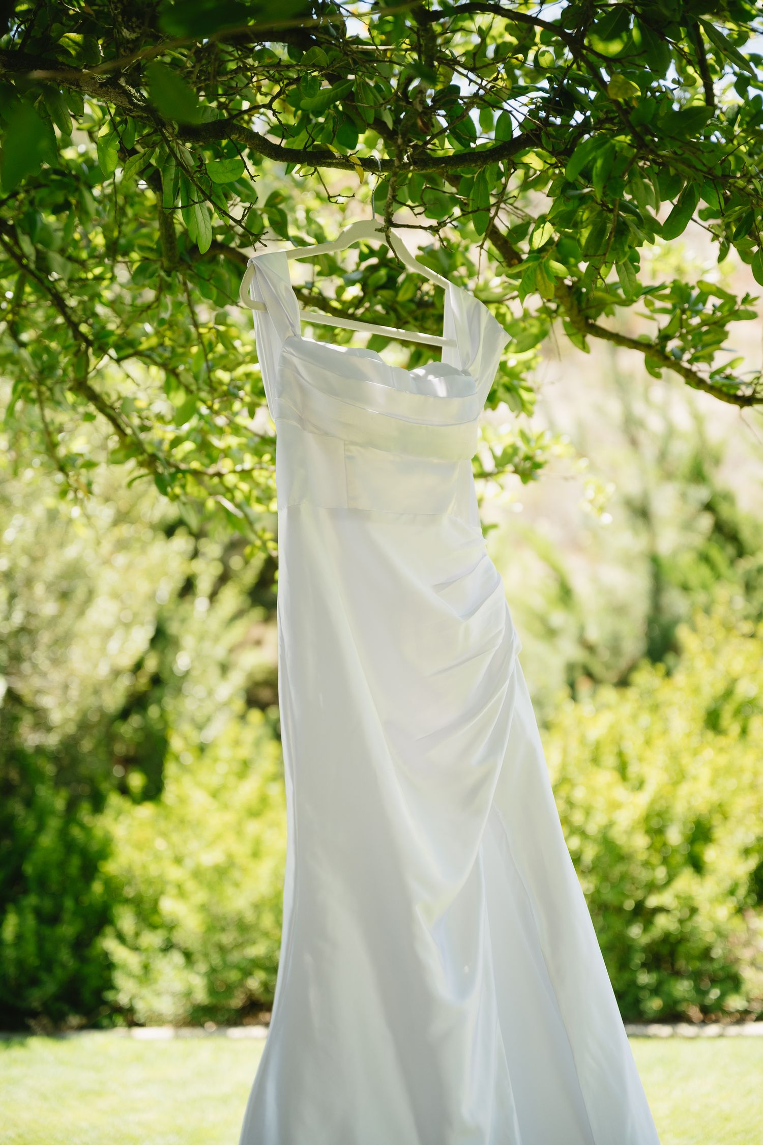 Photographer capturing a white wedding dress hanging from a tree branch in a lush green garden on a sunny day.