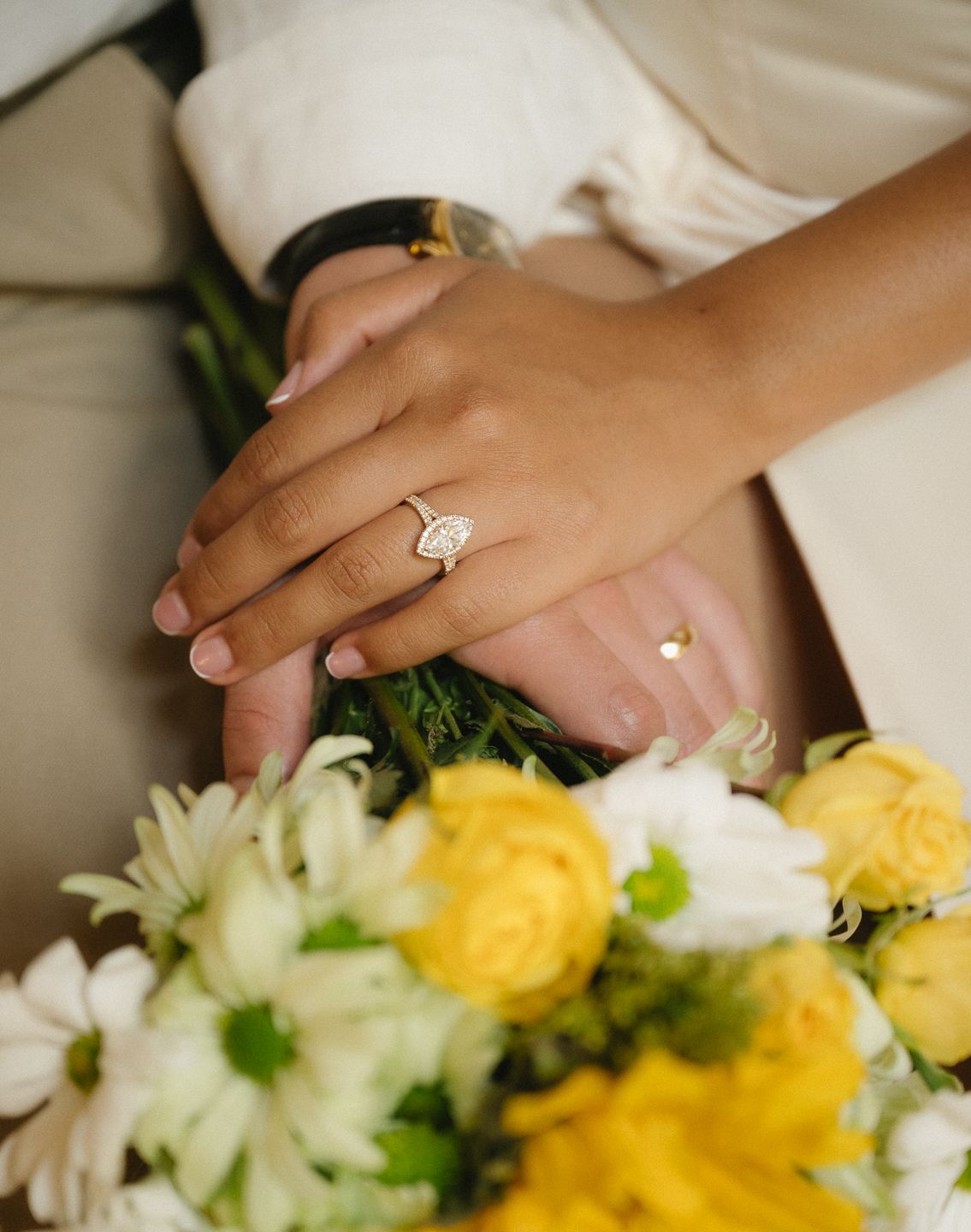 Close-up of bride's hand with elegant diamond halo engagement ring resting on colorful yellow and white wedding bouquet.