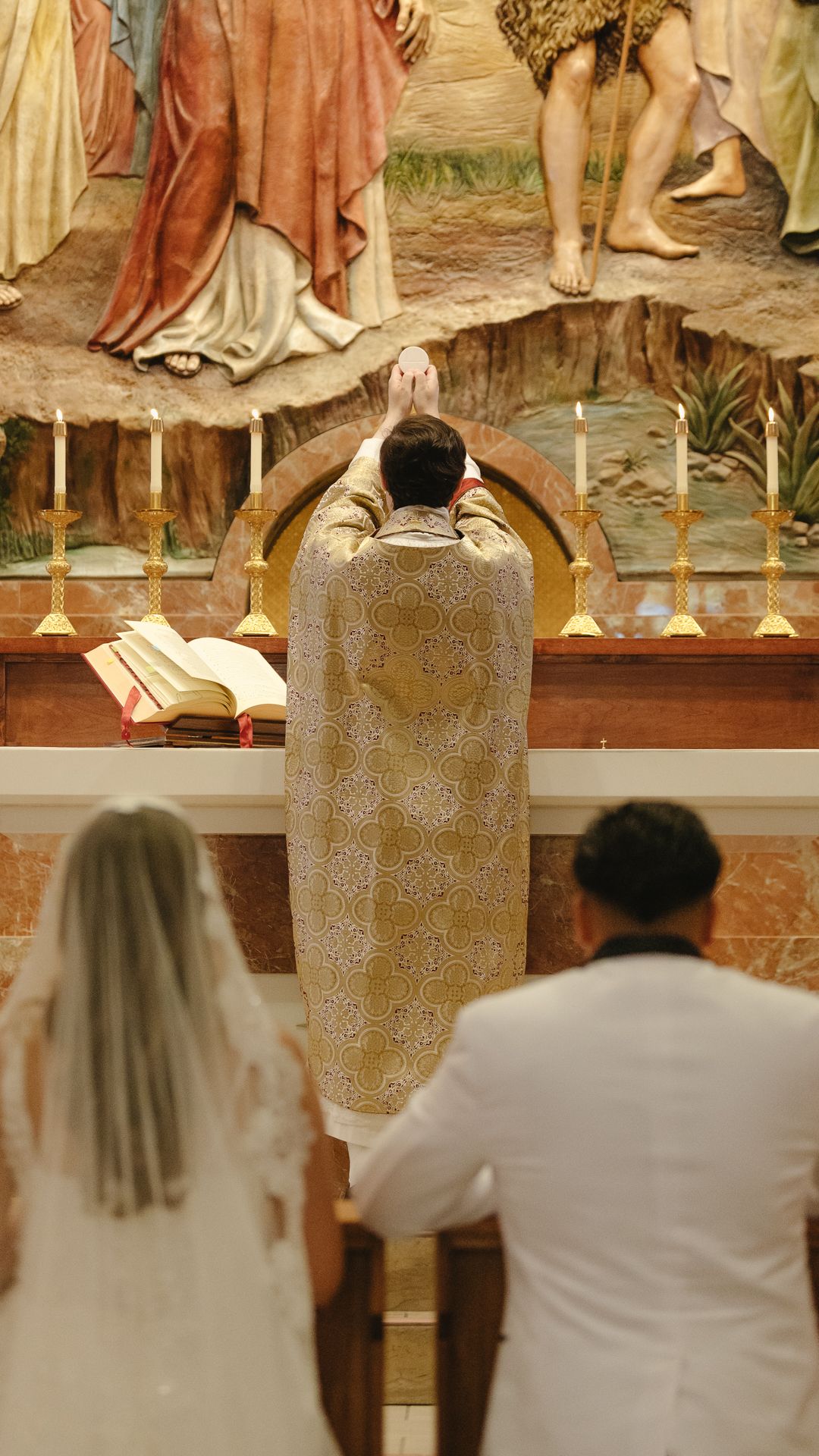 Catholic priest raises the Eucharist at a wedding ceremony altar, with bride and groom kneeling before ornate religious artwork.