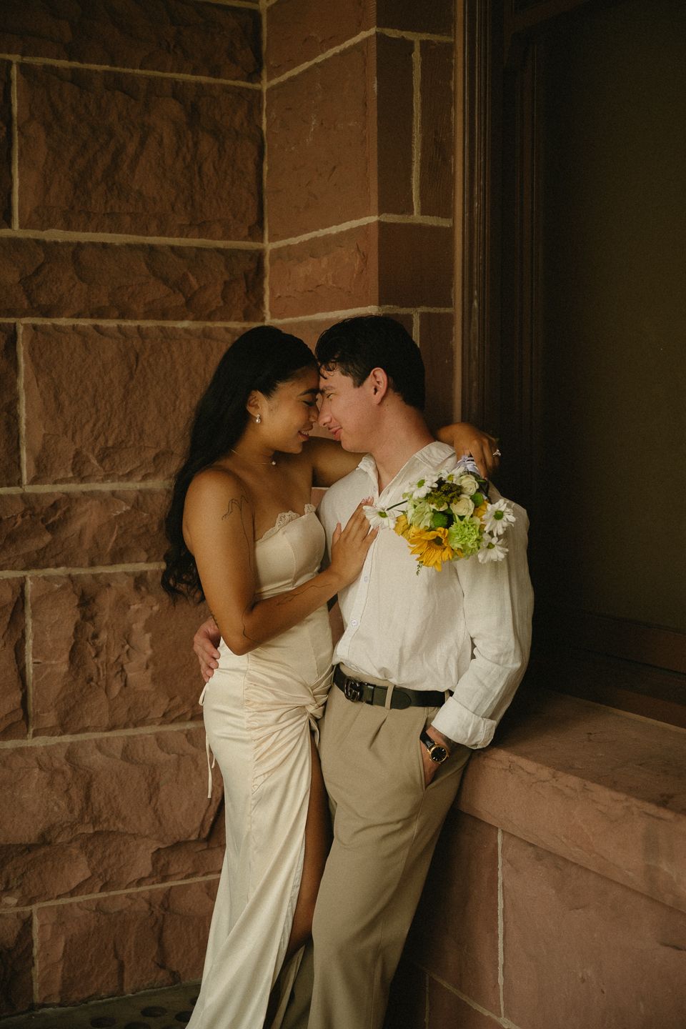 Bride and groom share a romantic moment leaning against a stone wall, bride holding a colorful bouquet.