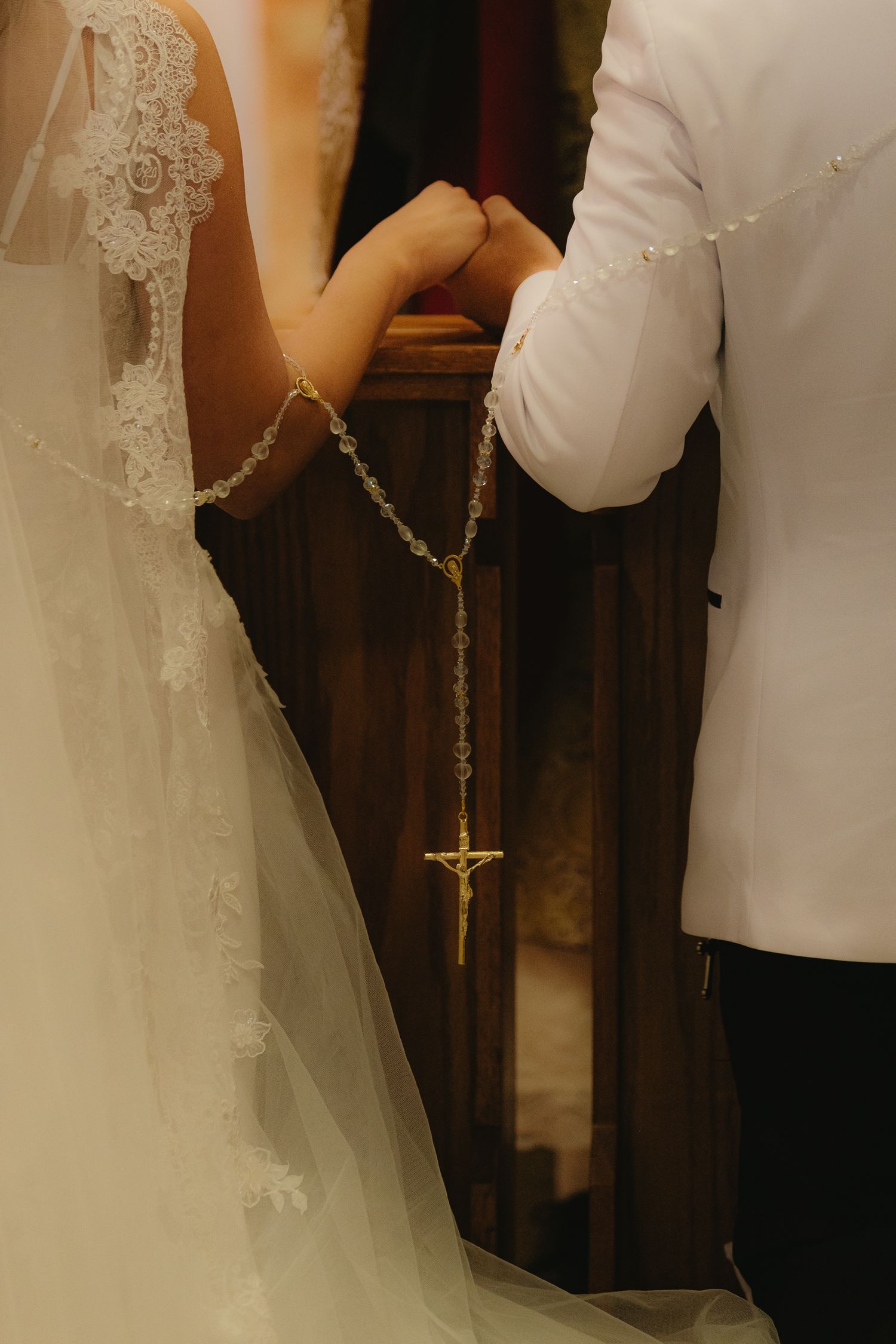 Bride and groom holding hands with a gold rosary cross during a church wedding ceremony.