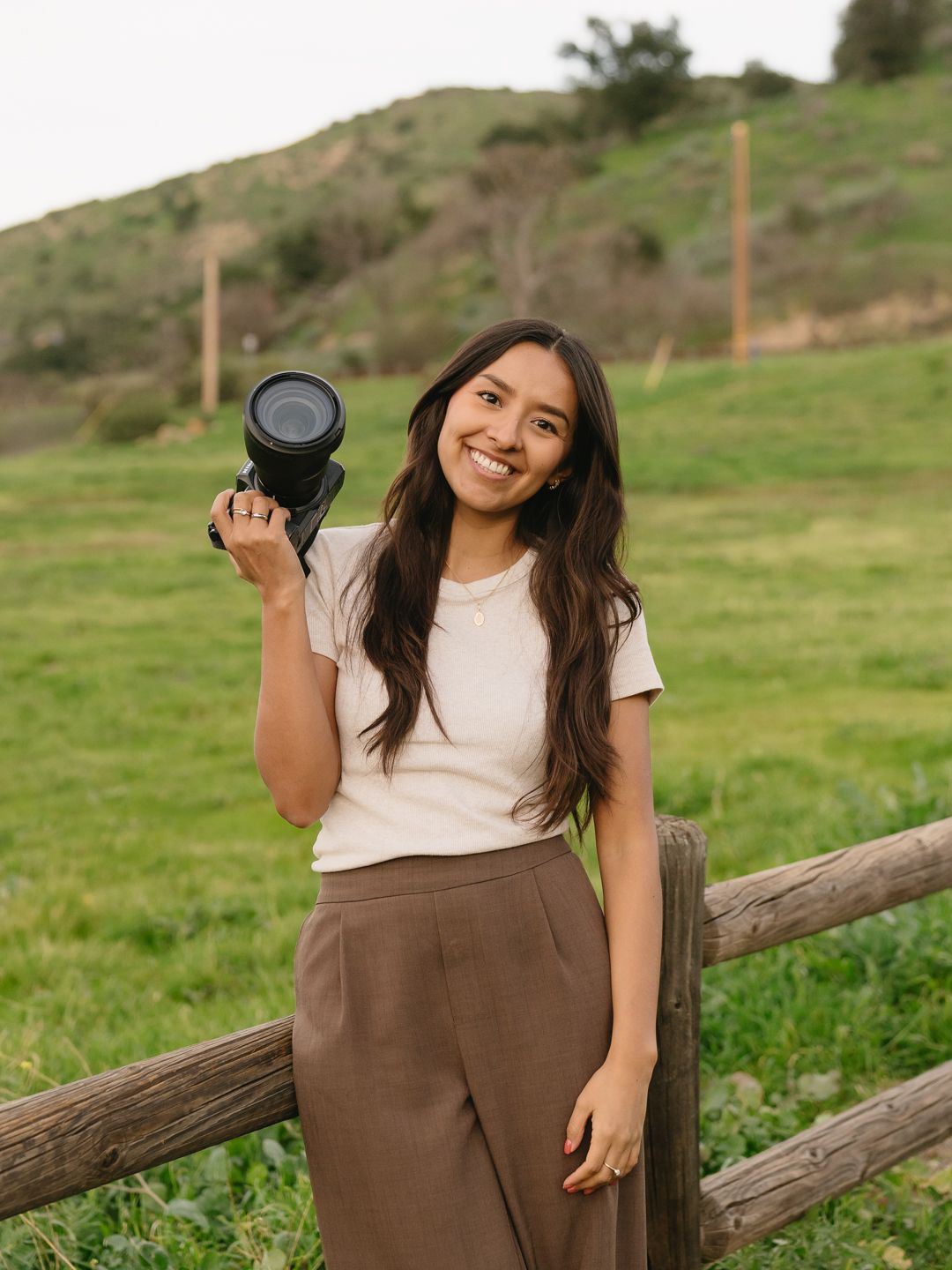 Smiling woman holding a camera outdoors, leaning on a wooden fence with green hills in the background.