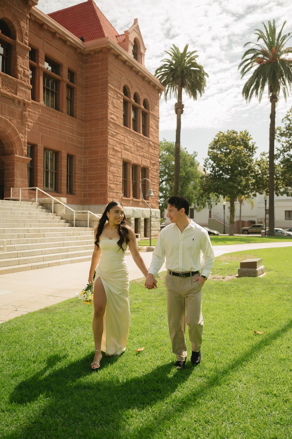 A couple in wedding attire holds hands walking on green grass beside a red brick building with palm trees.