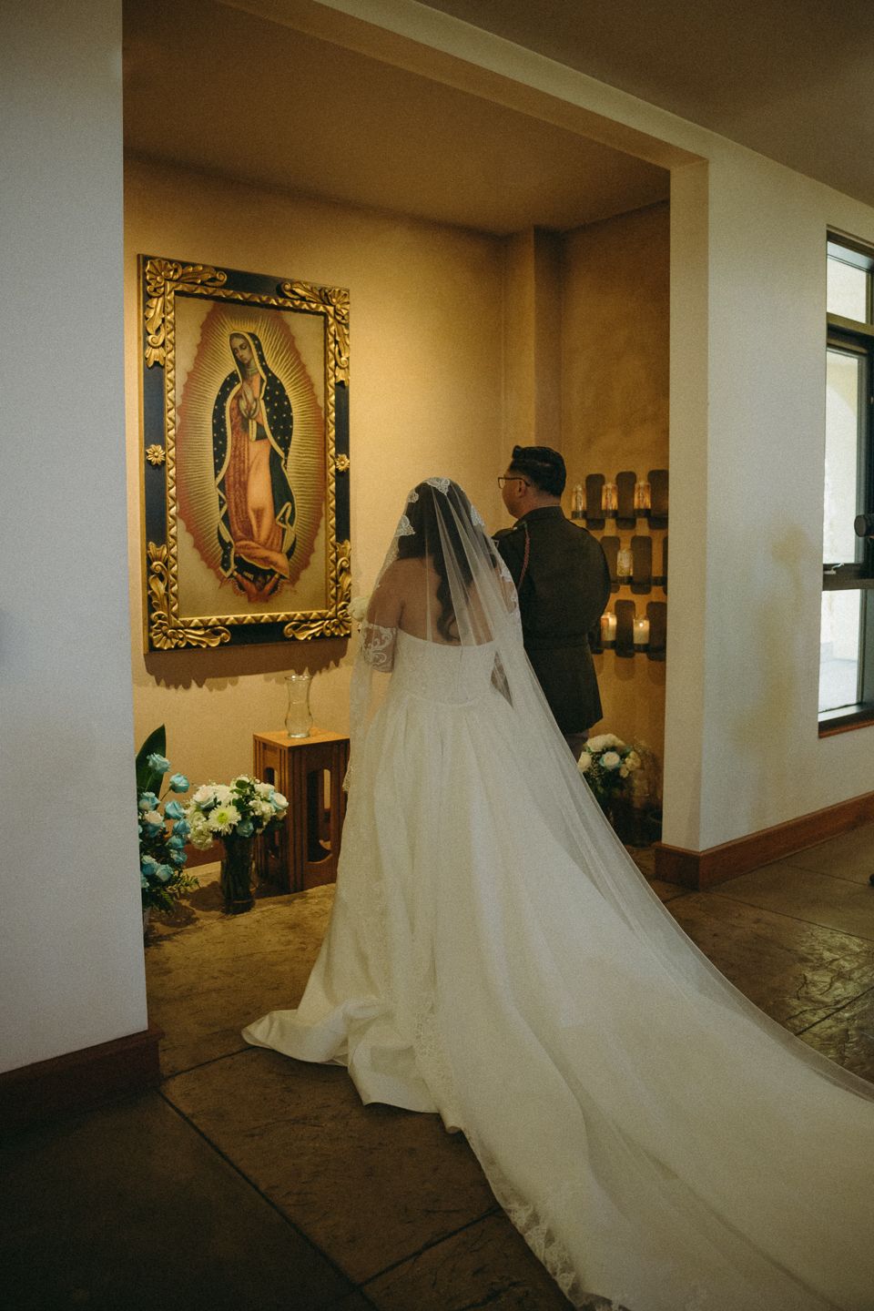 Bride in white gown and veil stands before Our Lady of Guadalupe painting in a chapel during wedding ceremony.