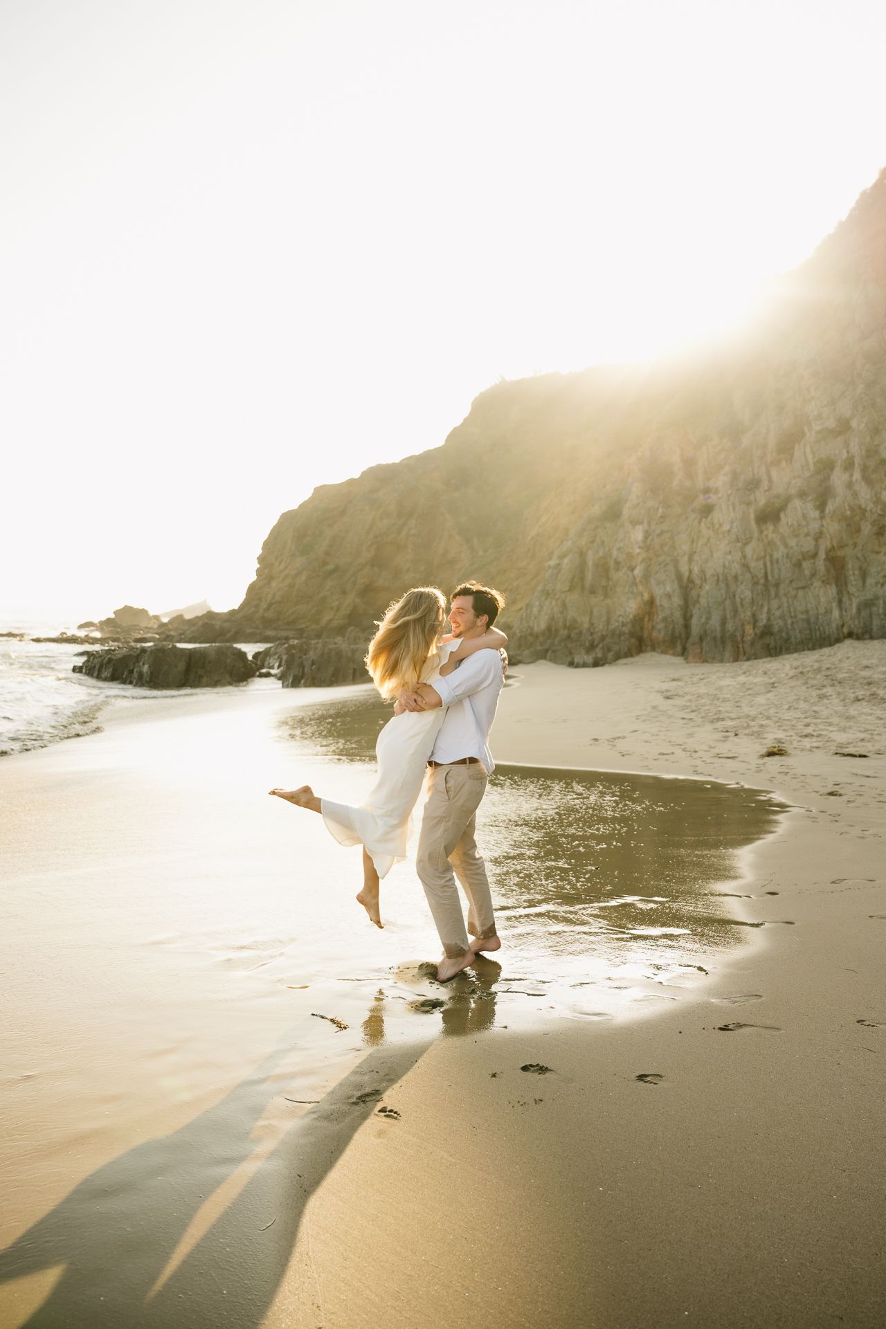 Couple embracing on a sunlit beach with golden light and rocky cliffs in the background.
