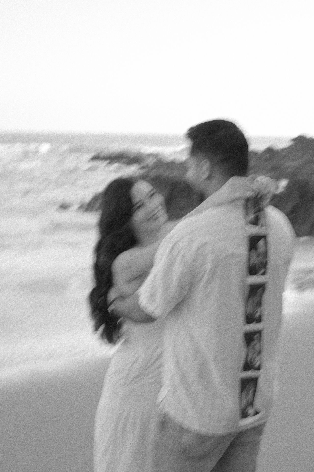 Black and white photo of a couple embracing on a beach, sharing a romantic moment with ocean waves in the background.