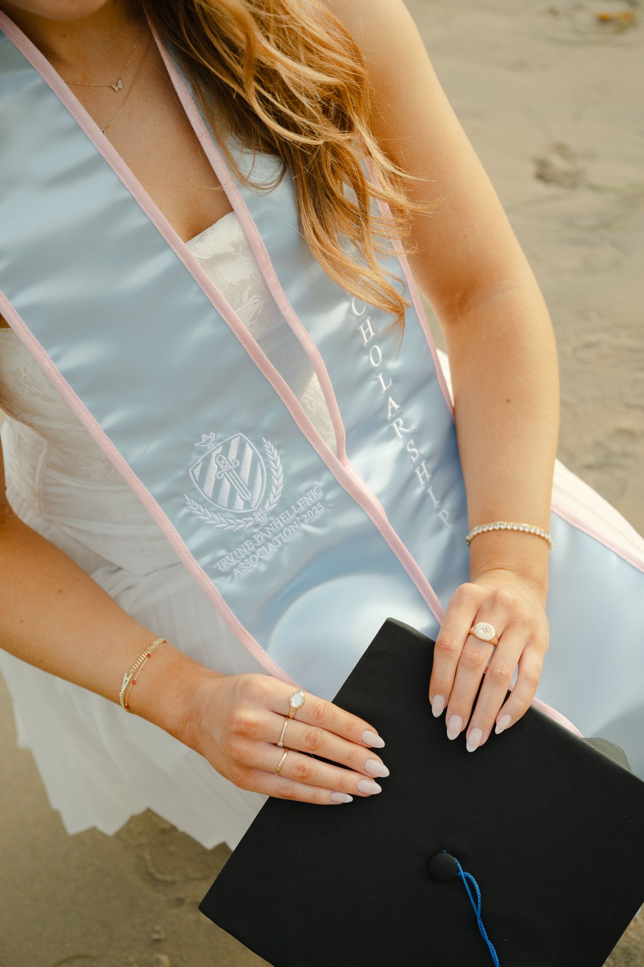 Graduate in white dress holds black cap, wearing light blue satin sash, gold bracelet, and rings on sandy beach.