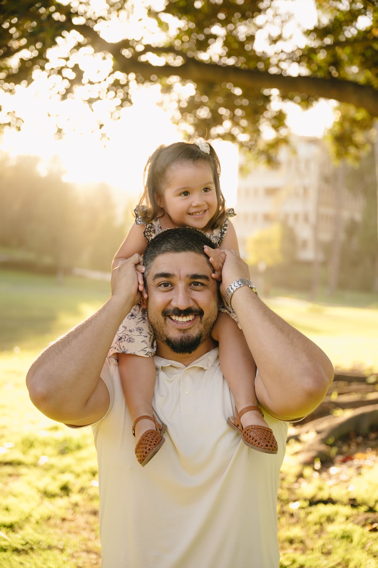 A smiling man carries a happy toddler girl on his shoulders in a sunlit park during golden hour.