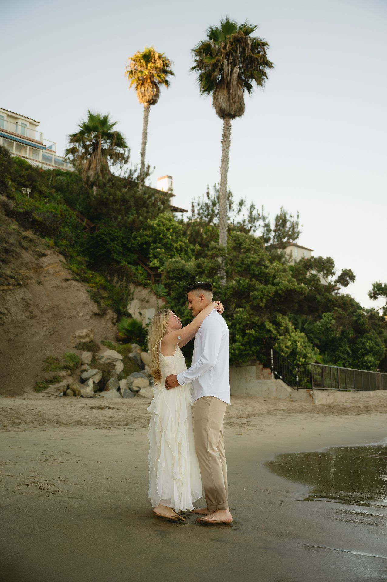 Couple dancing barefoot on a sandy beach with palm trees and coastal cliffs at sunset.