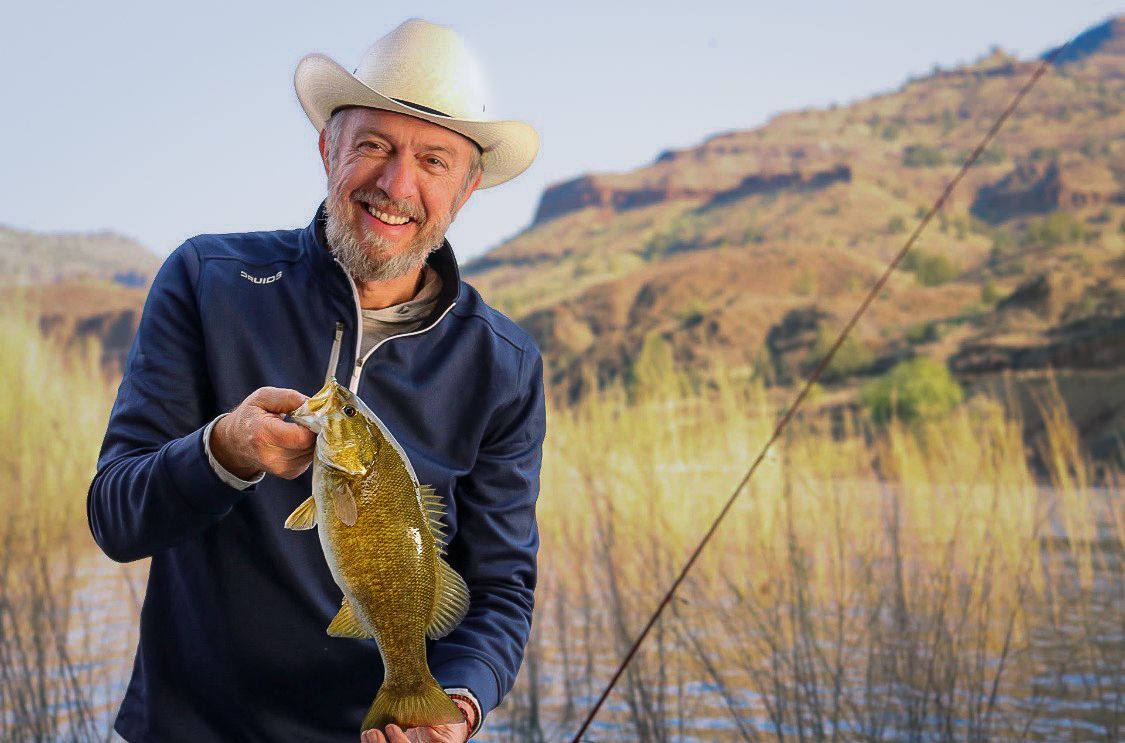 A smiling man in a cowboy hat proudly holds a smallmouth bass by a scenic river.