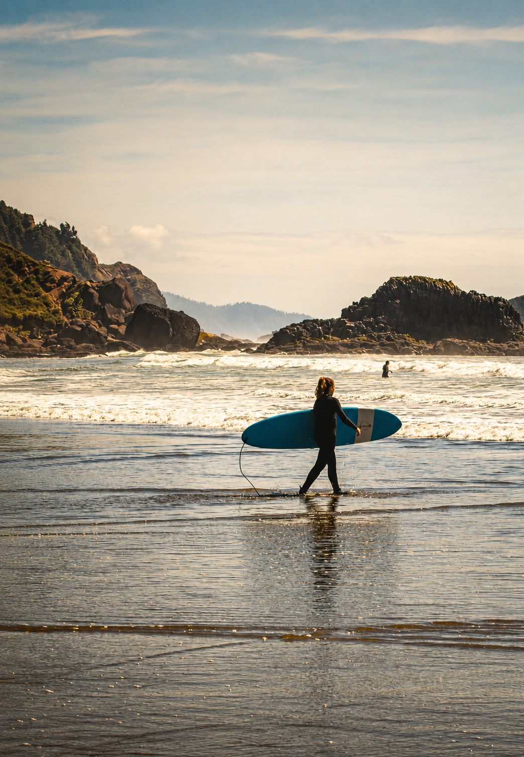 A surfer carries a blue longboard along a scenic rocky beach at golden hour.