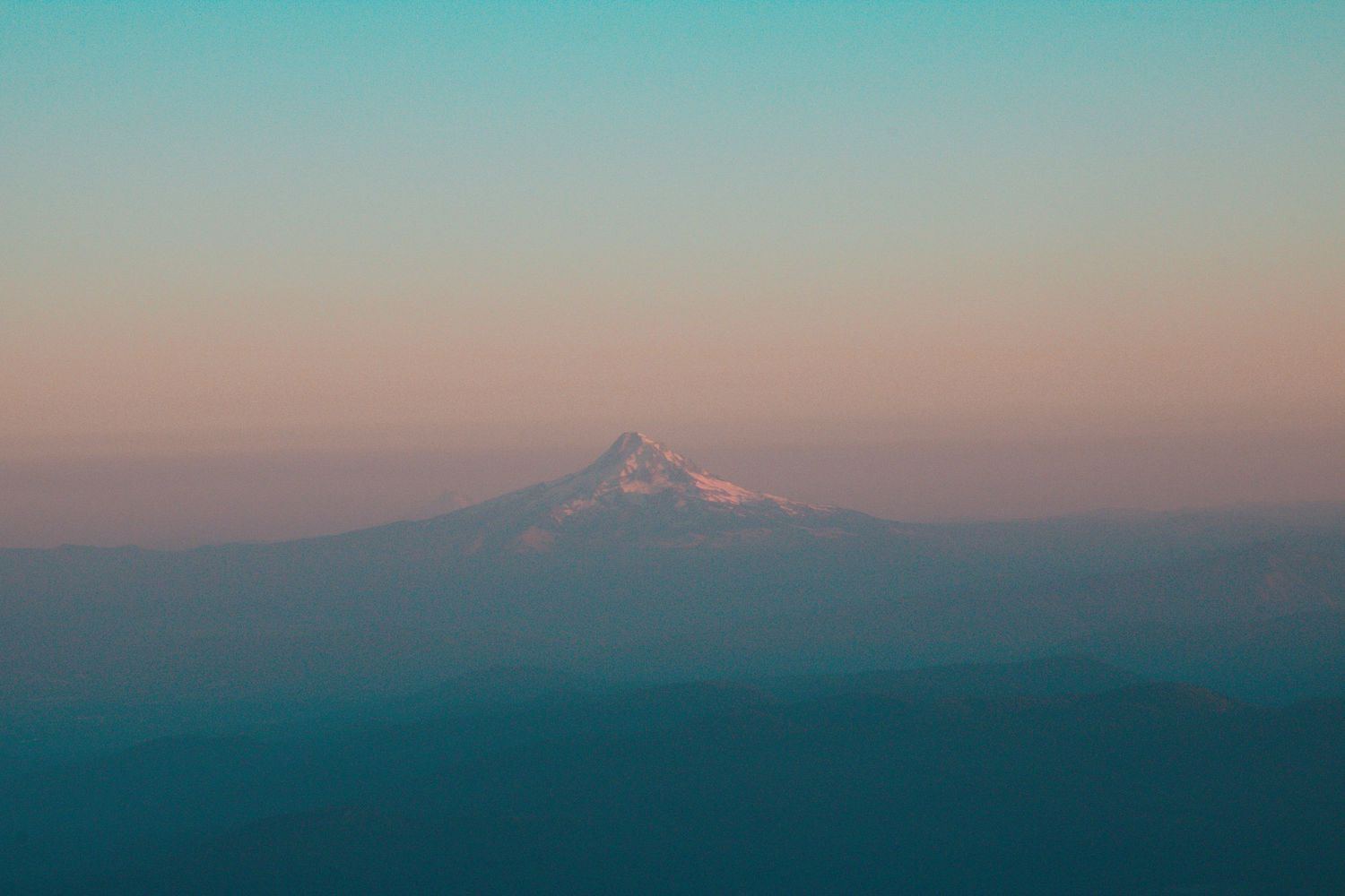 Snow-capped Mount Hood rising above layered blue mountain ridges during a colorful pastel sunset.
