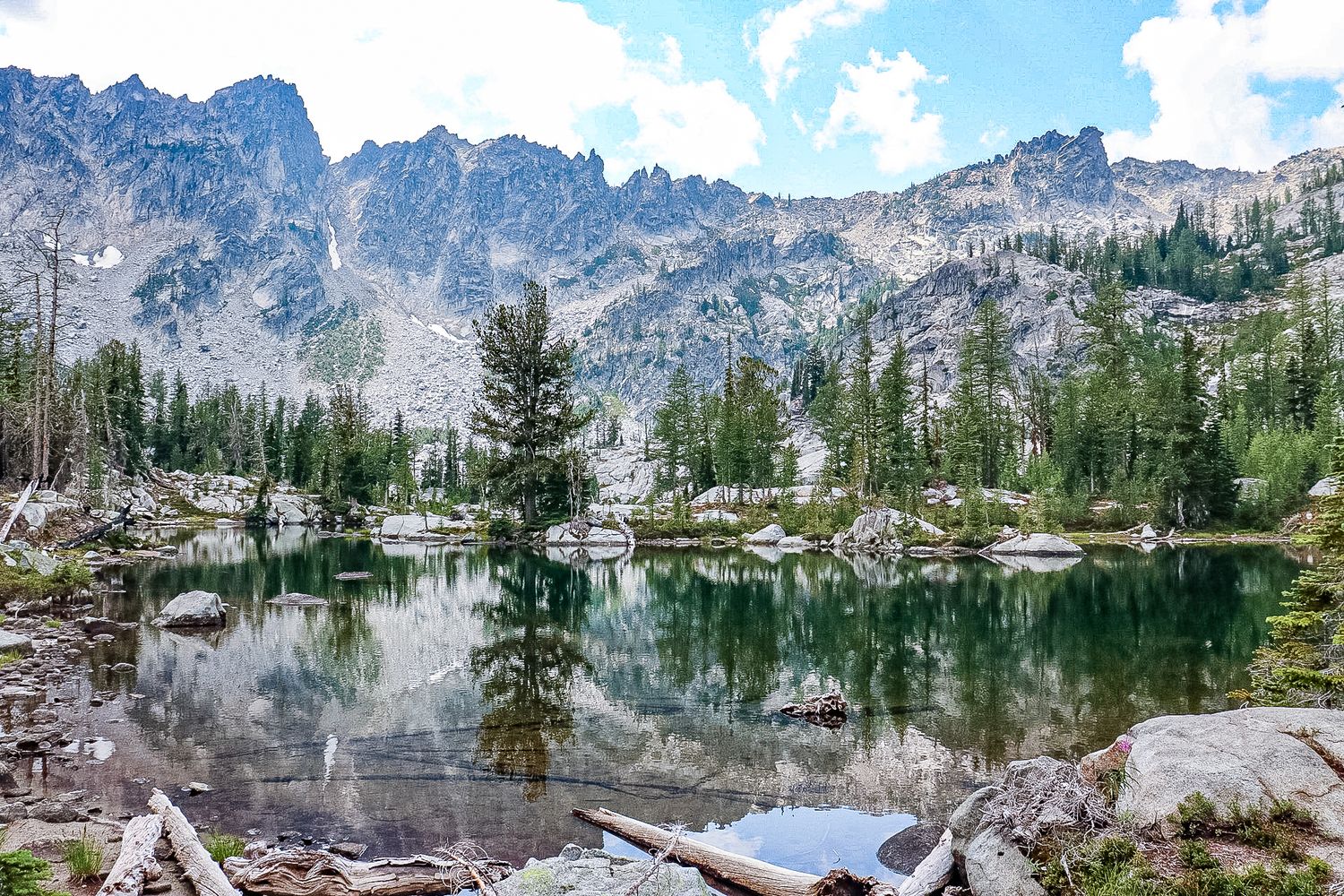 Crystal-clear alpine lake reflecting pine trees and jagged granite peaks in a wilderness setting.