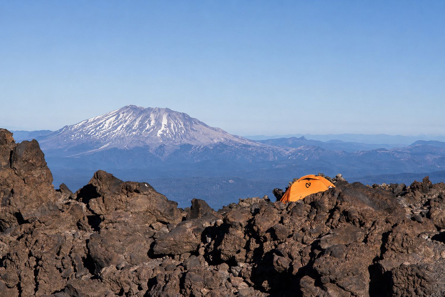 Orange camping tent nestled among volcanic rocks with a snow-capped mountain peak in the background.