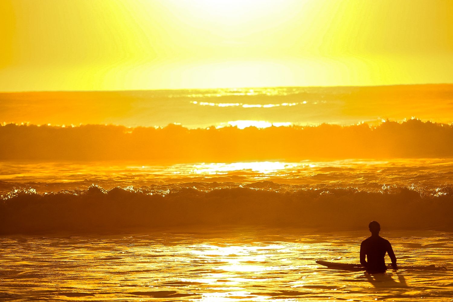 Silhouette of a surfer holding a board in golden ocean waves at sunset.