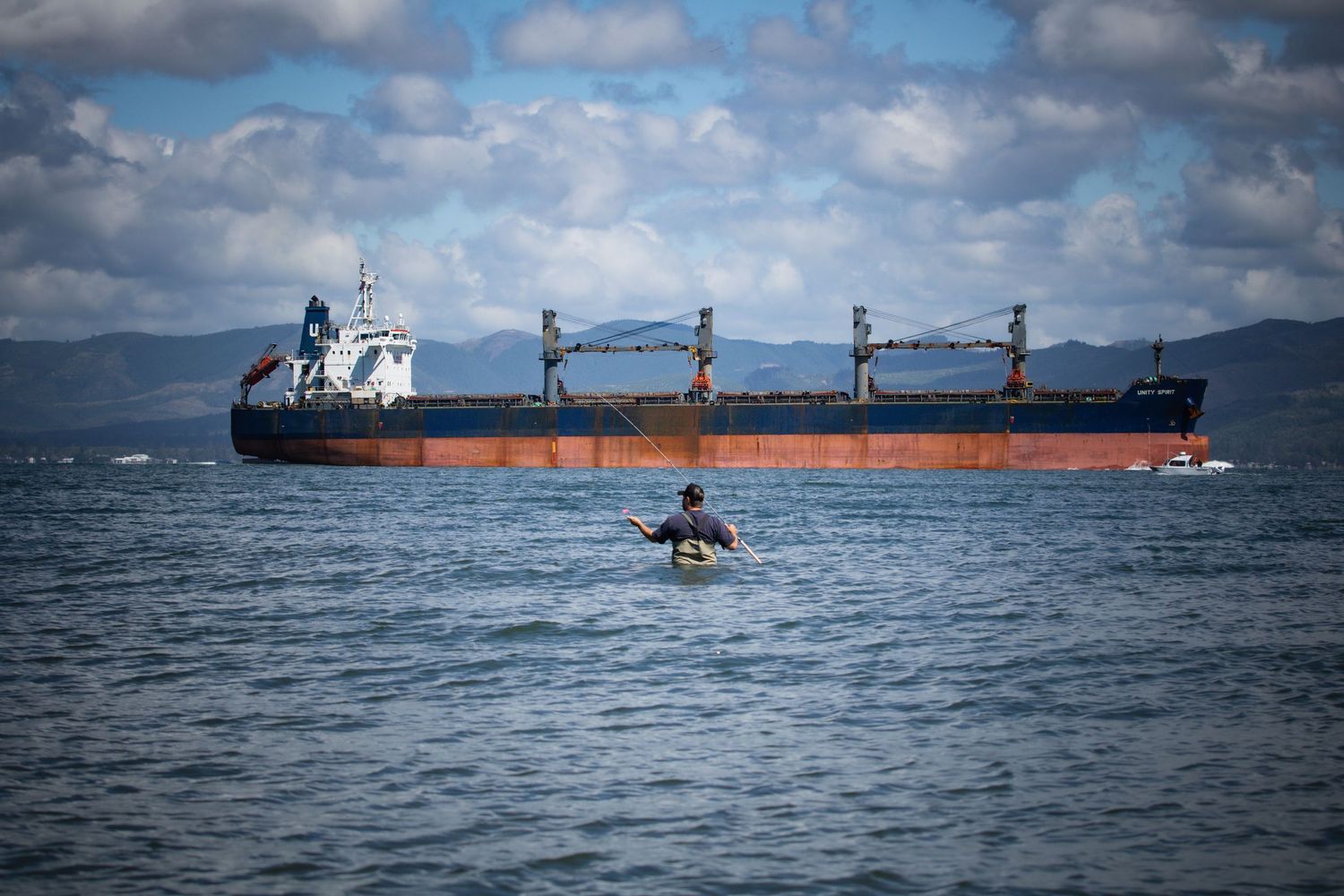 A kayaker paddles near a large cargo ship on calm ocean waters with mountains in the background.