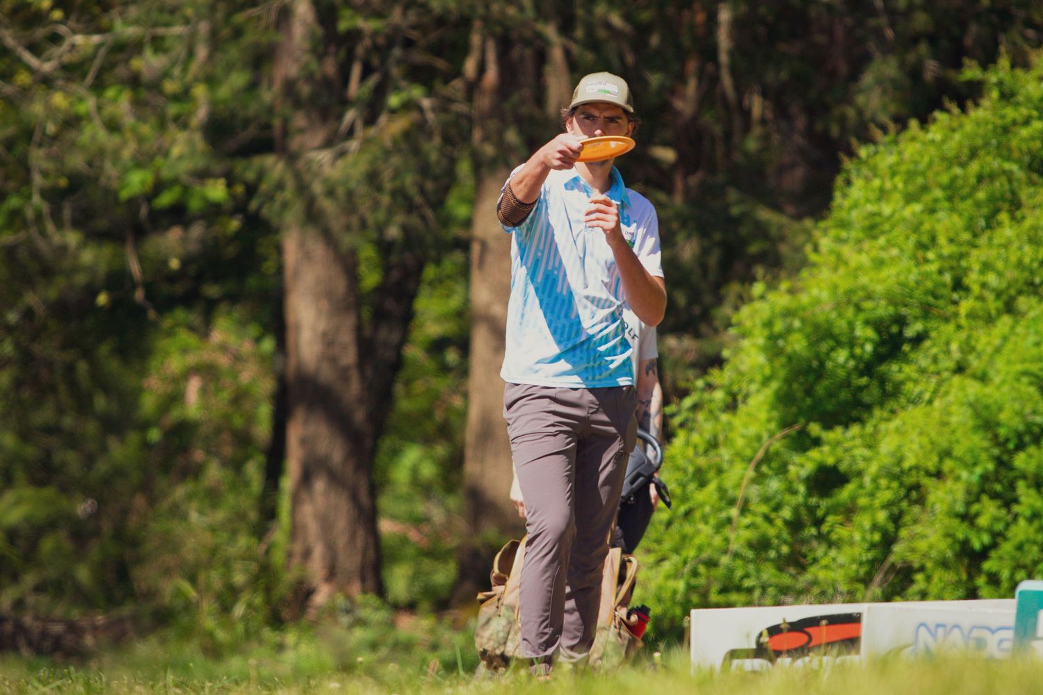 A man in a hat and blue shirt plays disc golf in a lush green outdoor course.