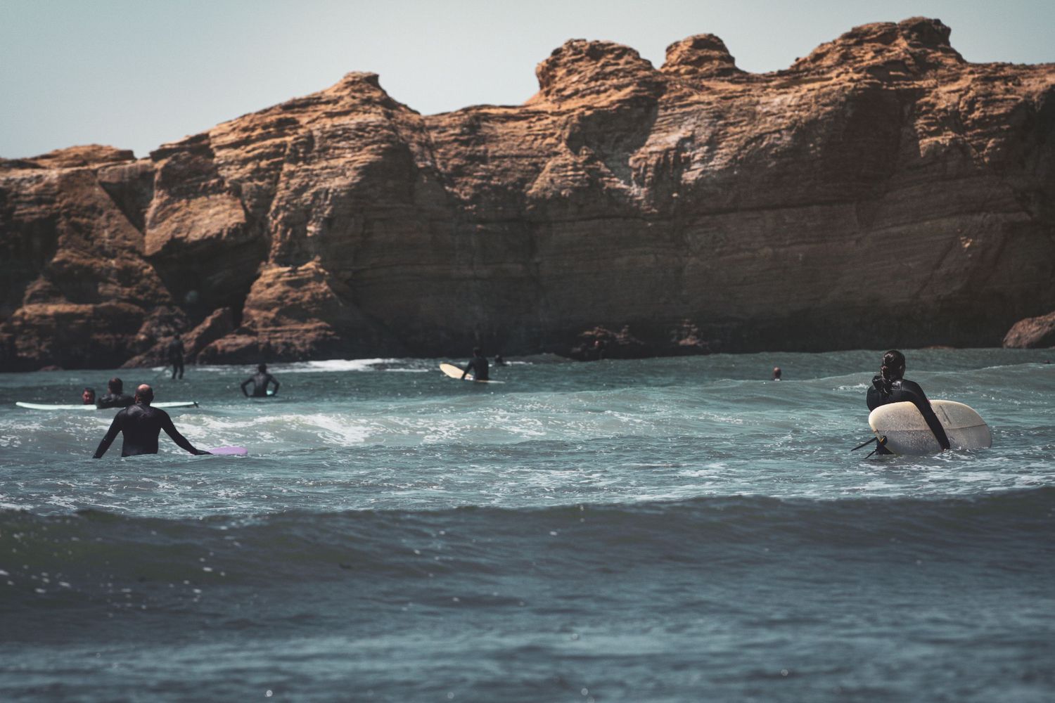 A group of surfers paddle and carry boards in the surf near rugged rocky coastal cliffs.