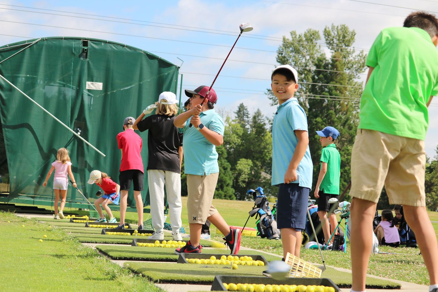 Children practice golf swings at an outdoor driving range during a youth golf lesson on a sunny day.