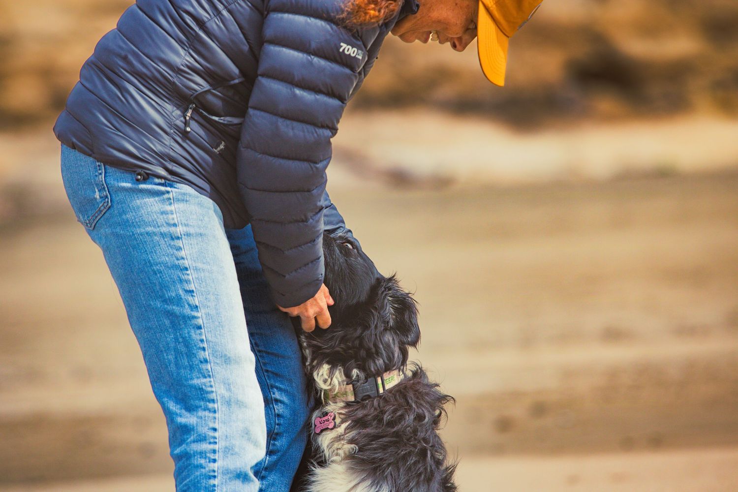 Person in blue puffer jacket and yellow cap petting a fluffy dog outdoors on sandy terrain.