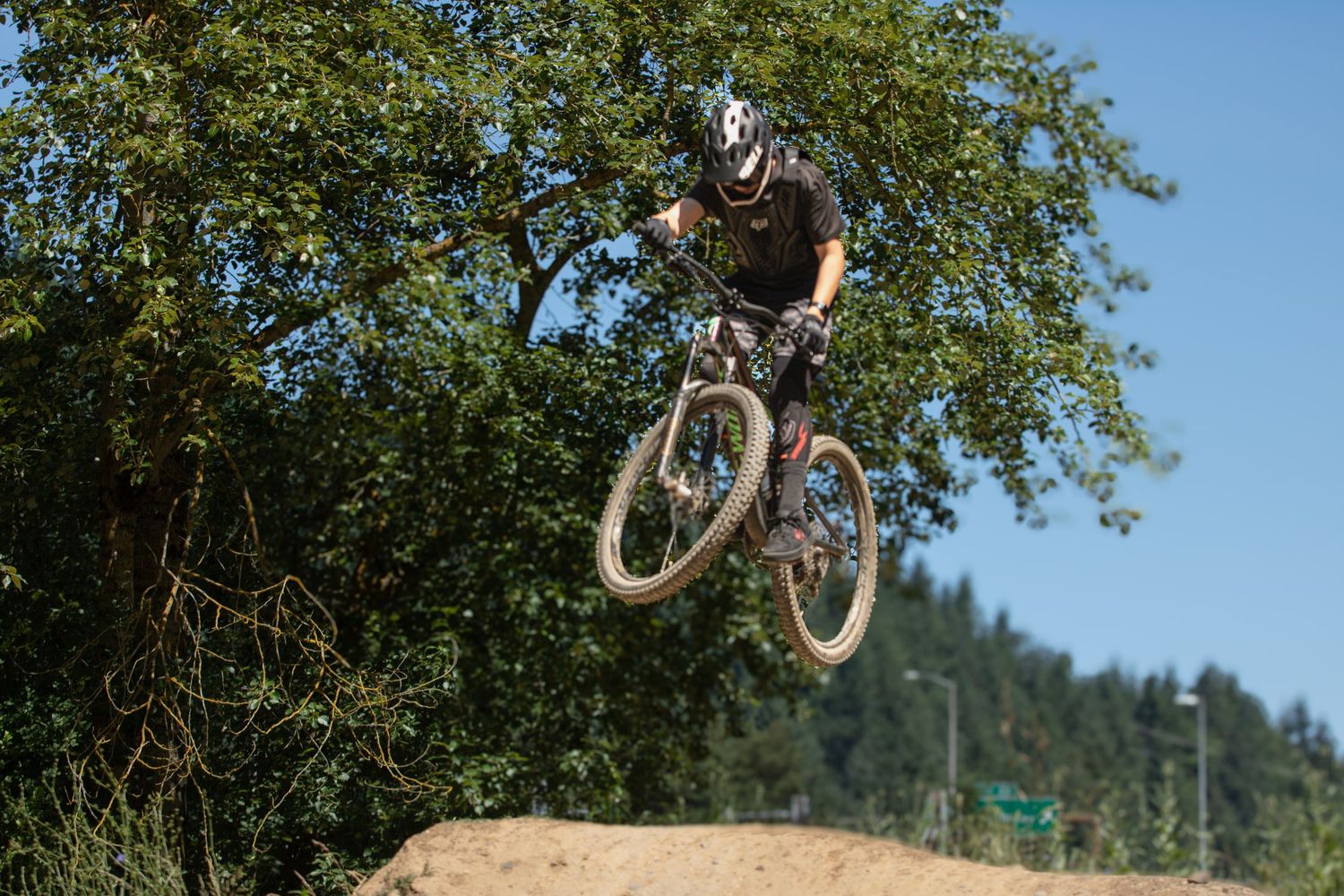 Mountain biker performing aerial jump trick over dirt mound against blue sky.