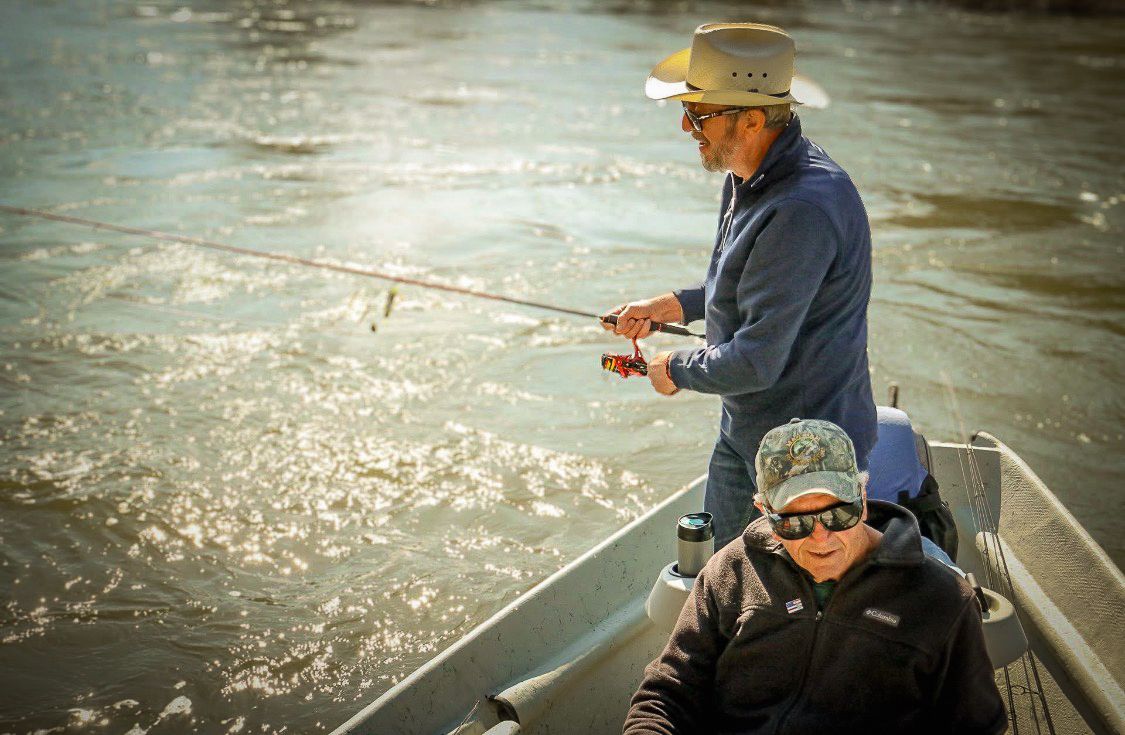 Two men fish from a small aluminum boat on a sparkling river, one standing casting a rod.
